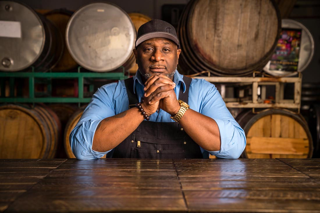 Bertony Faustin Oregons First Black Winemaker Abbey Creek Vineyard founder sitting at a wooden table in overalls in front of wine barrels