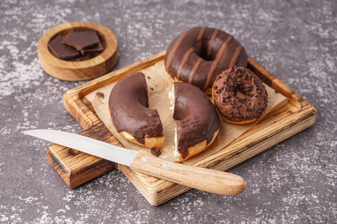A steak knife on a chopping block next to three chocolate donuts