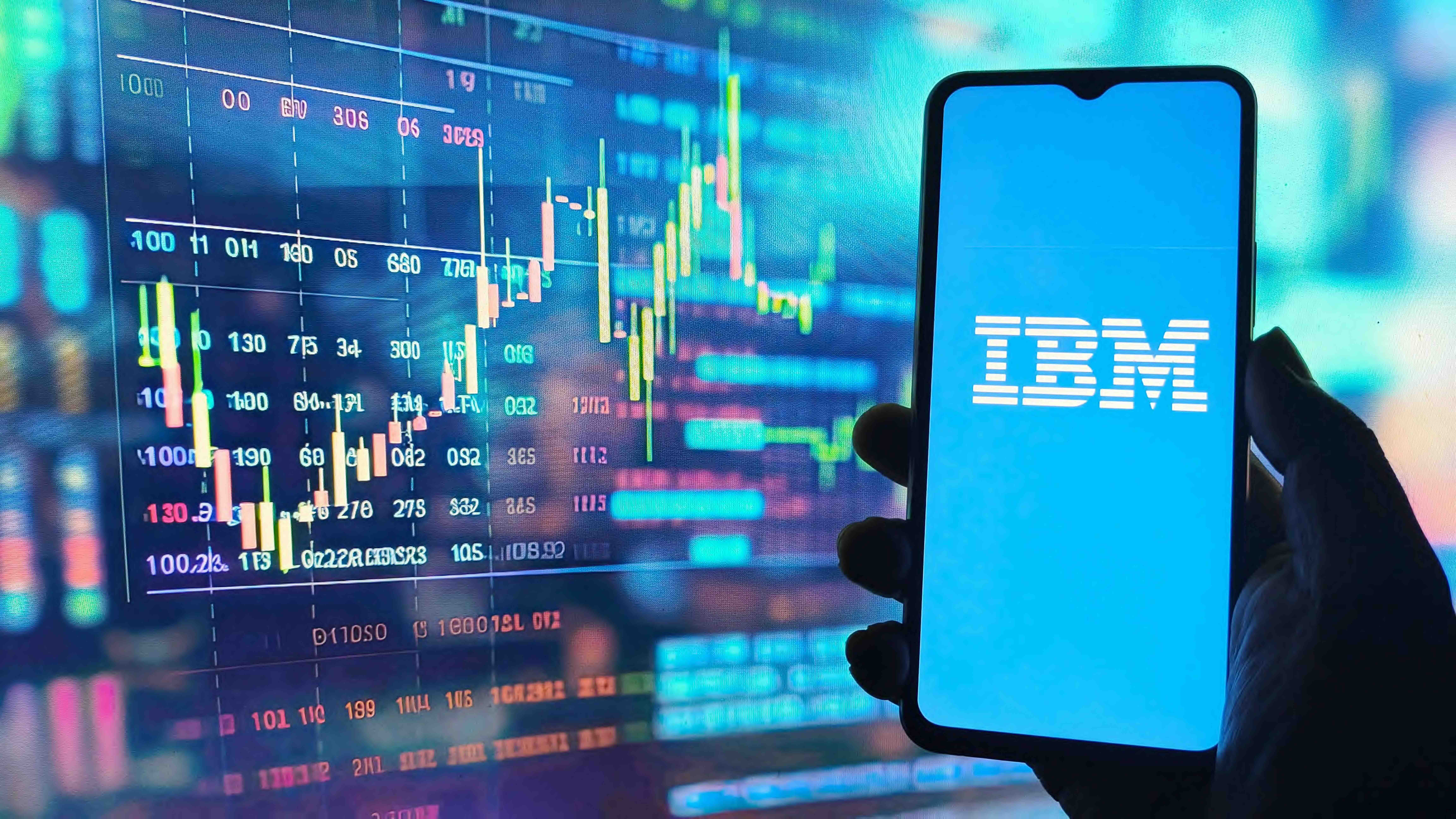 A stock market chart displaying declining candlestick data on a screen in the background, with a hand holding a smartphone displaying the IBM logo in the foreground.
