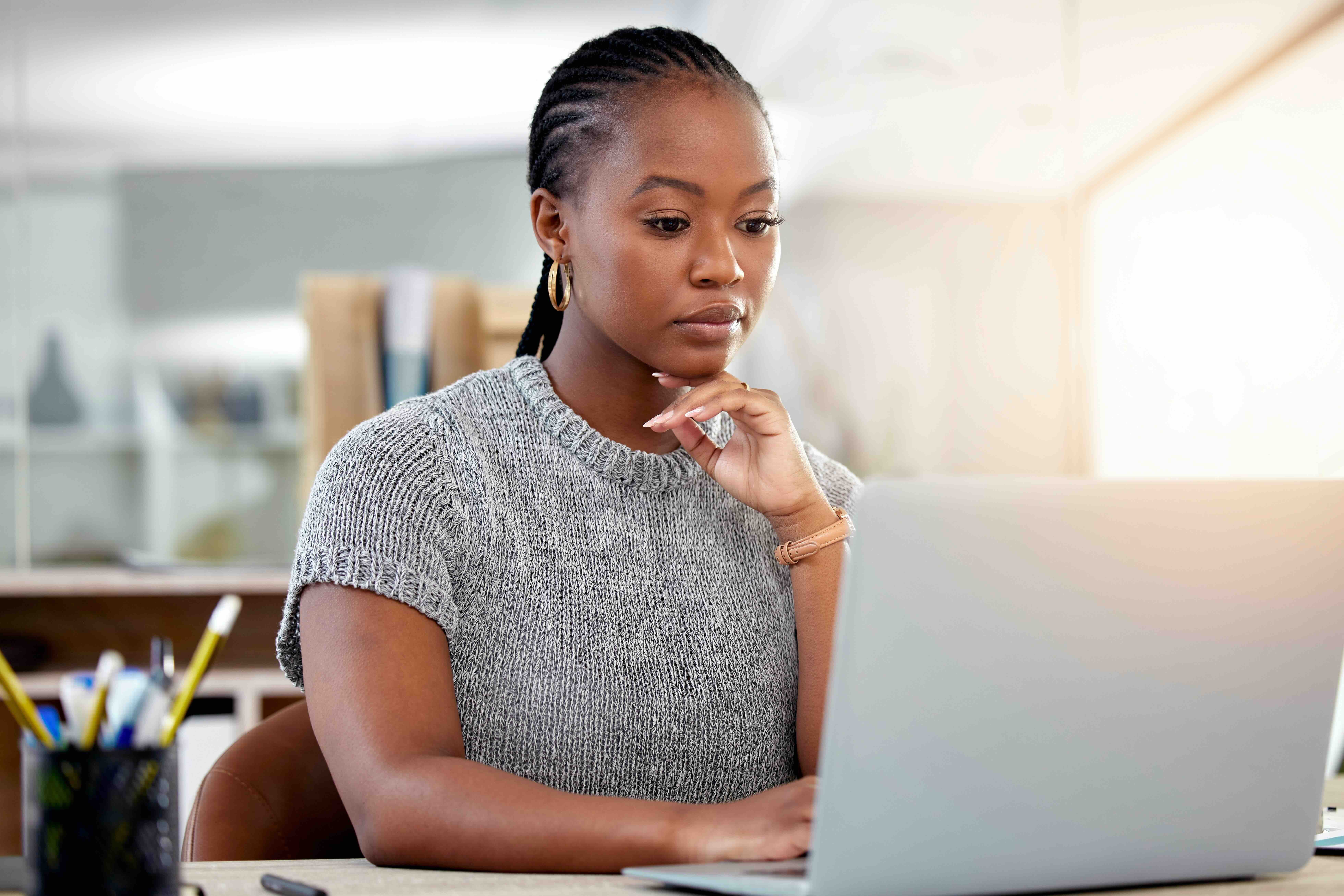 Woman in gray sweater concentrating while working on laptop at desk, appearing thoughtful about professional development