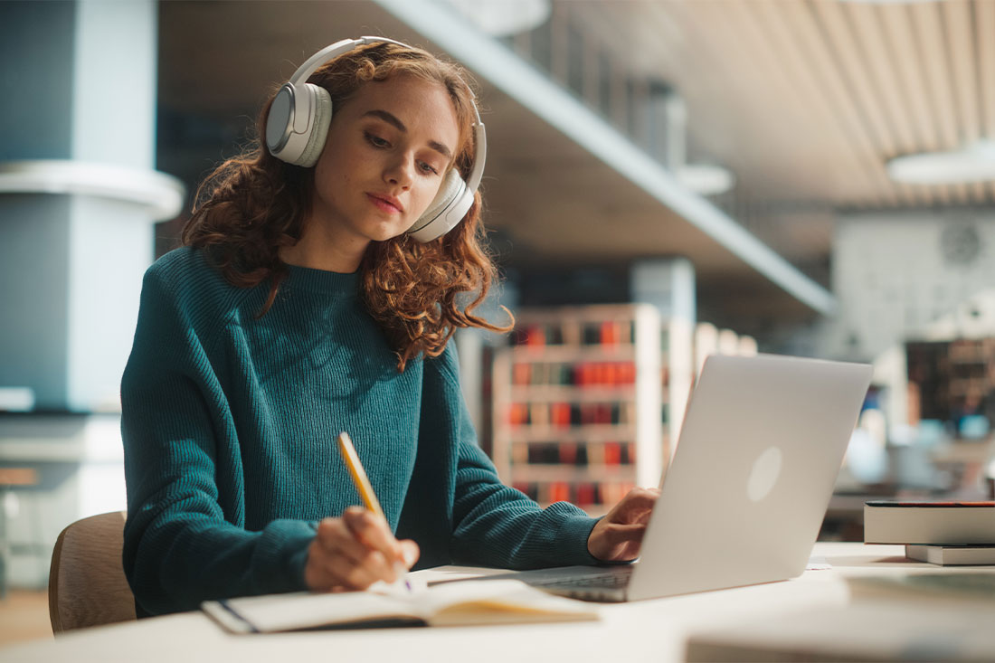 A young female student engages in academic research at a library desk with a laptop and headphones