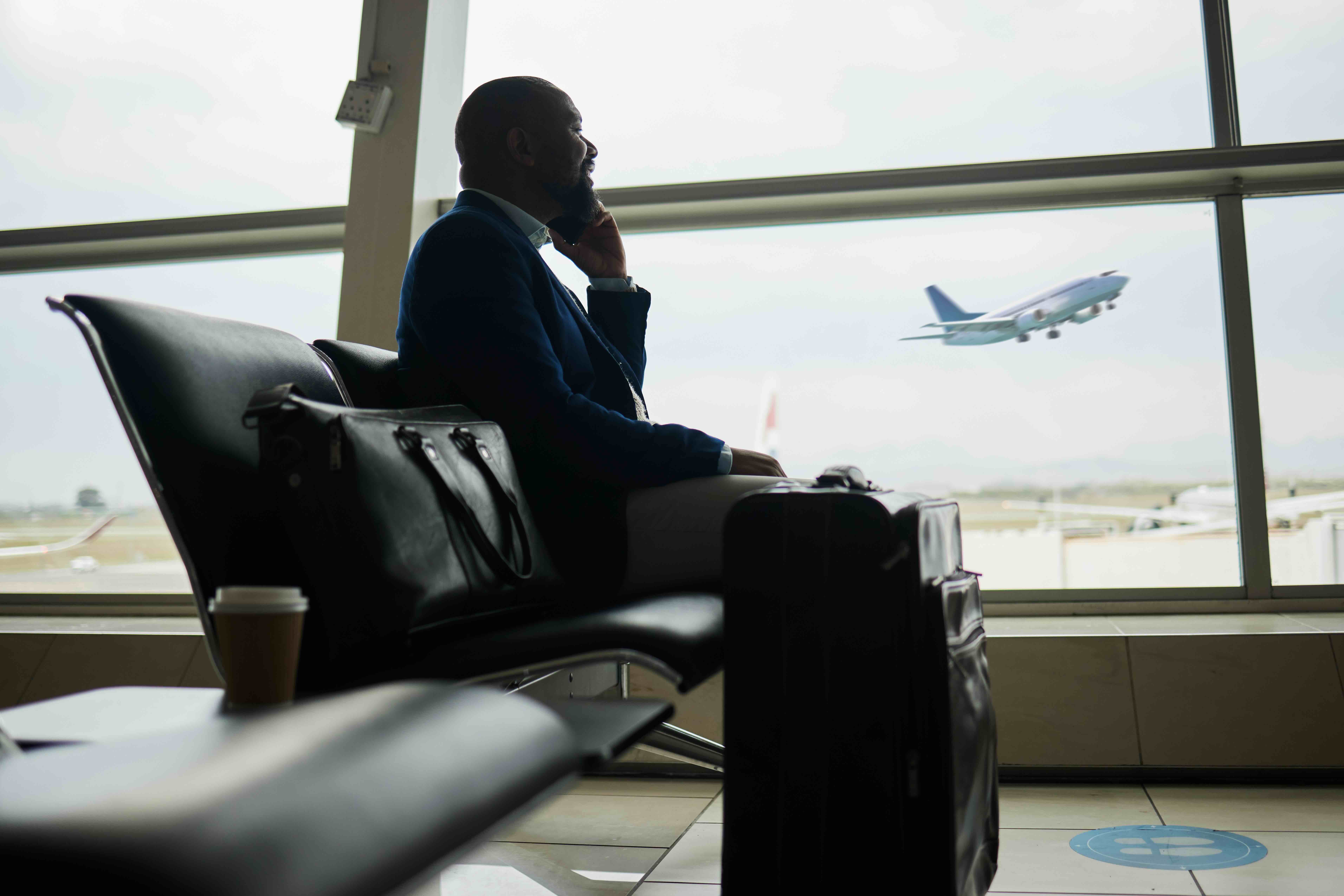 Business traveler in suit sits in airport terminal on phone with luggage as plane takes off in background