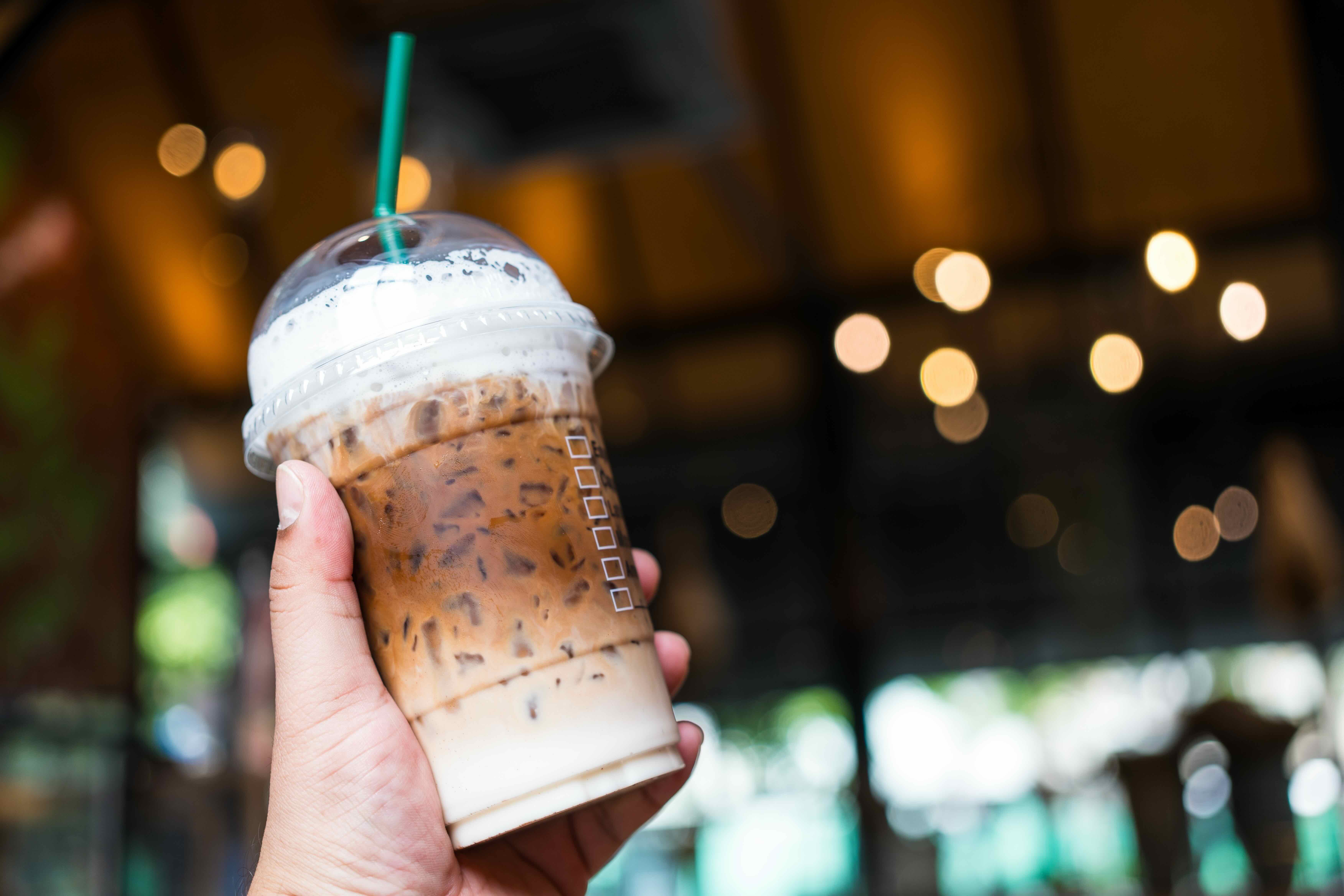 Hand holding iced coffee drink with whipped cream and straw against blurred café background with bokeh lights