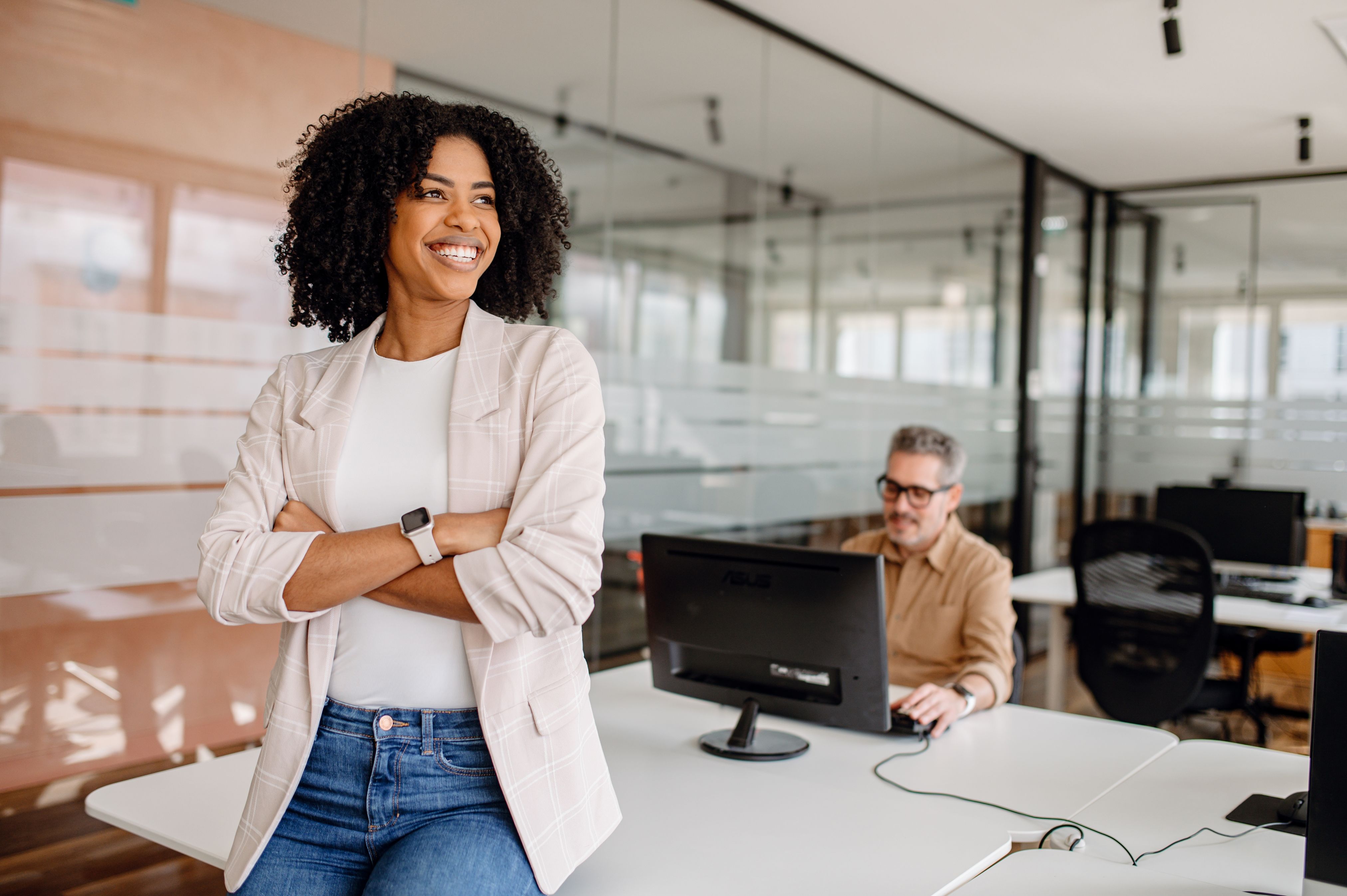 Professional woman with confident smile standing in modern office workspace with colleague at computer in background
