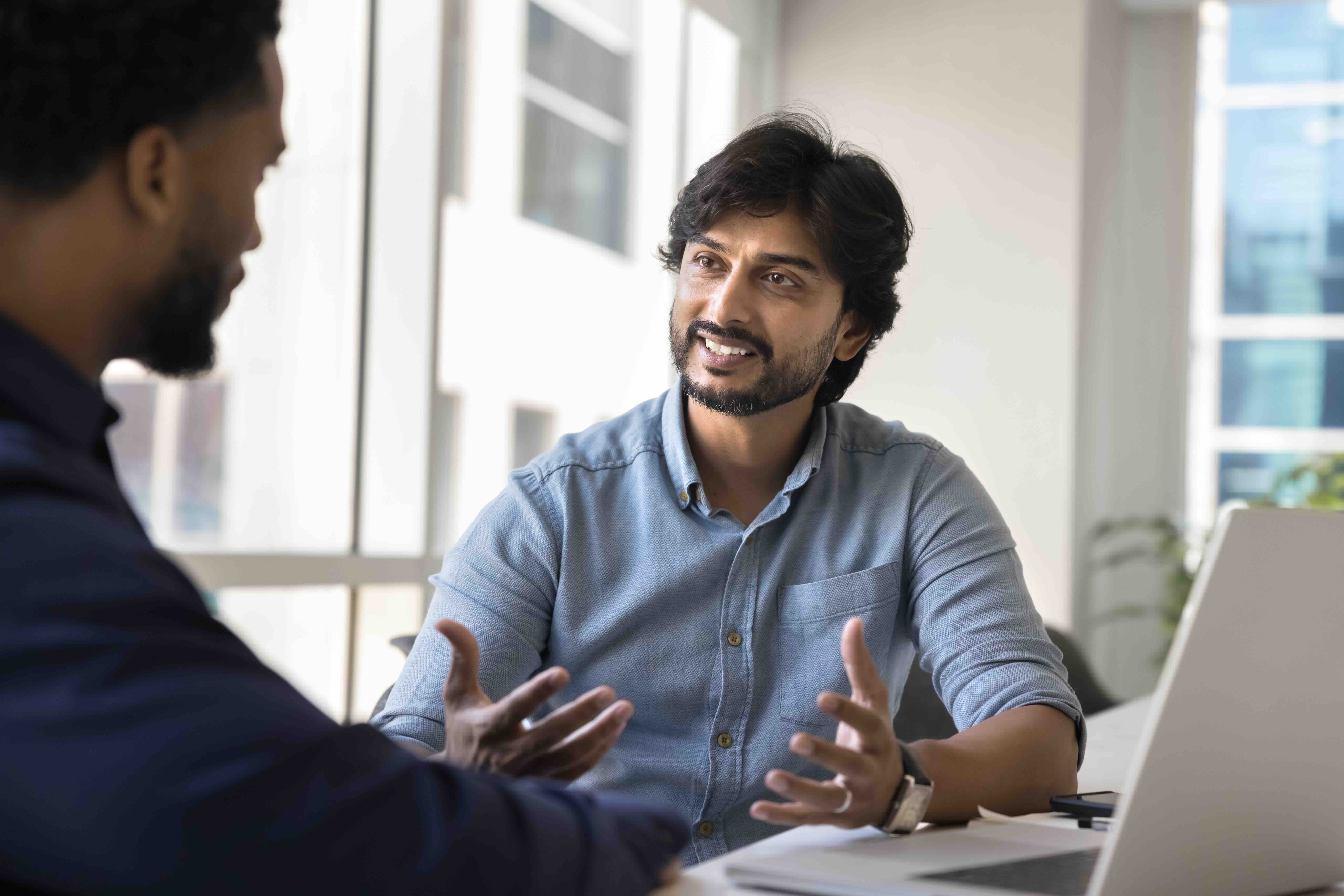 A smiling bearded man in a light blue shirt gestures openly as he talks with a client across a desk with a laptop in a bright office.