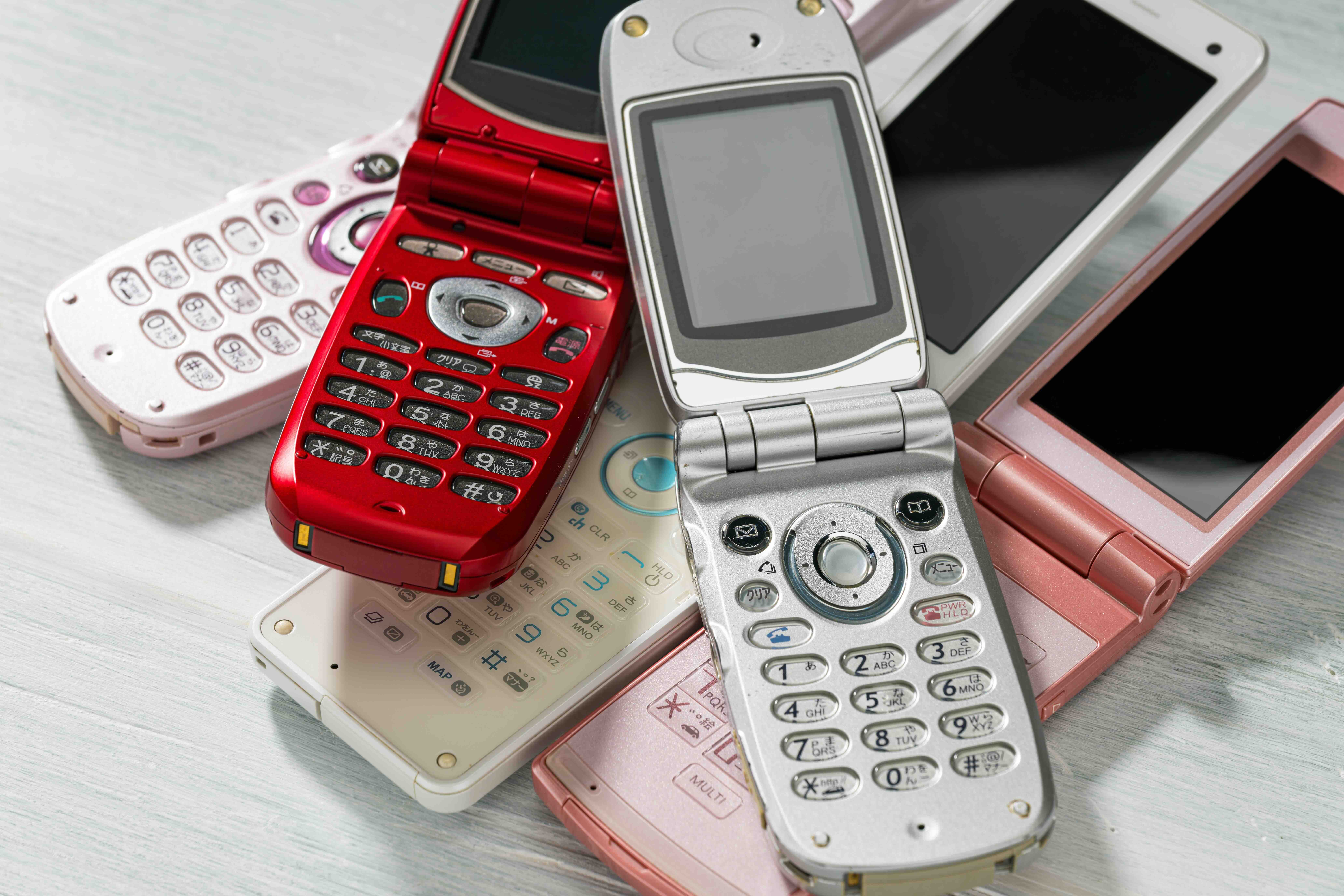 Pile of old flip phones in various colors — red, silver, pink, and white — stacked together on a light wooden surface.