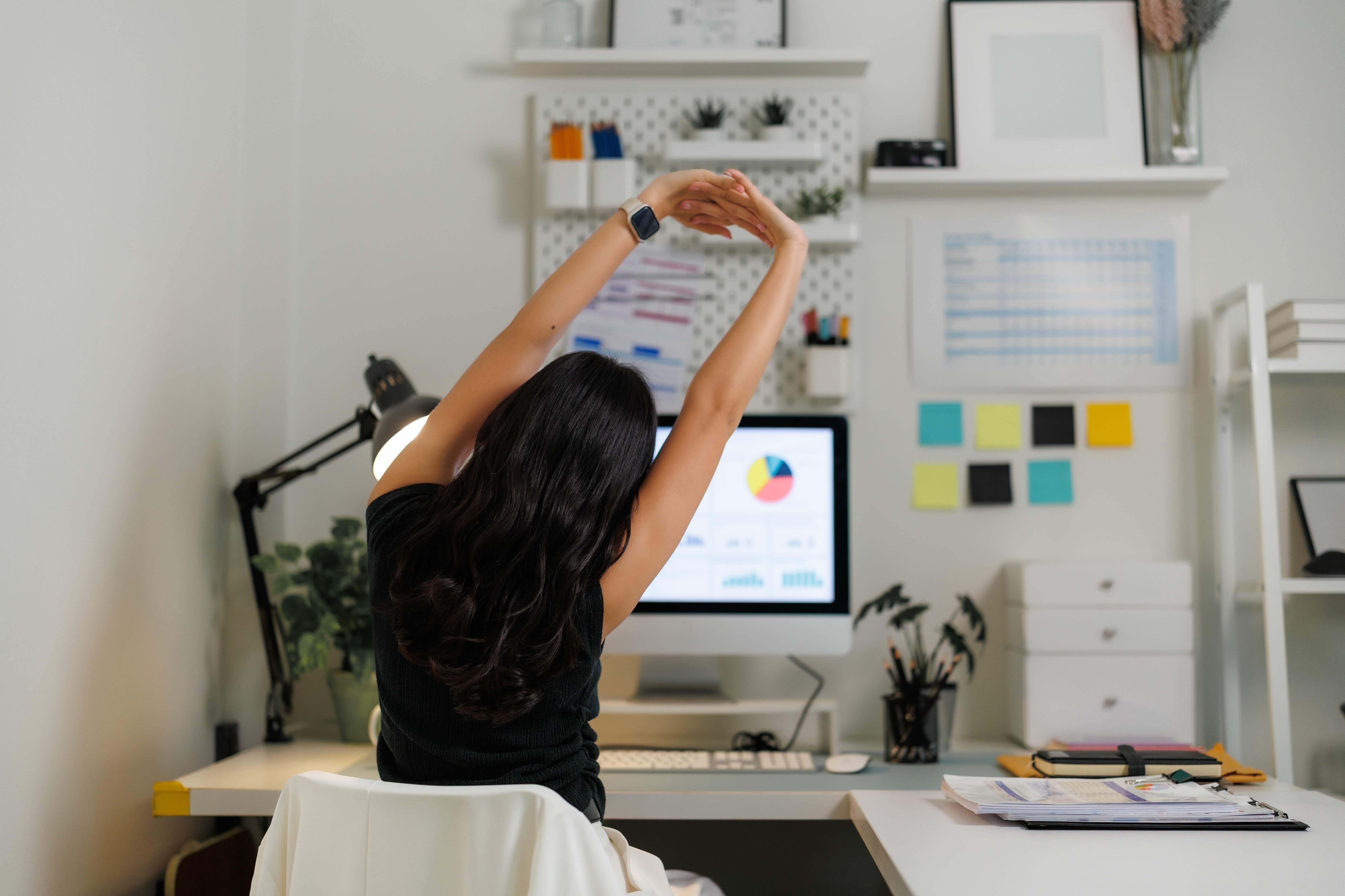Woman with long dark hair stretching arms overhead at organized home office desk with computer