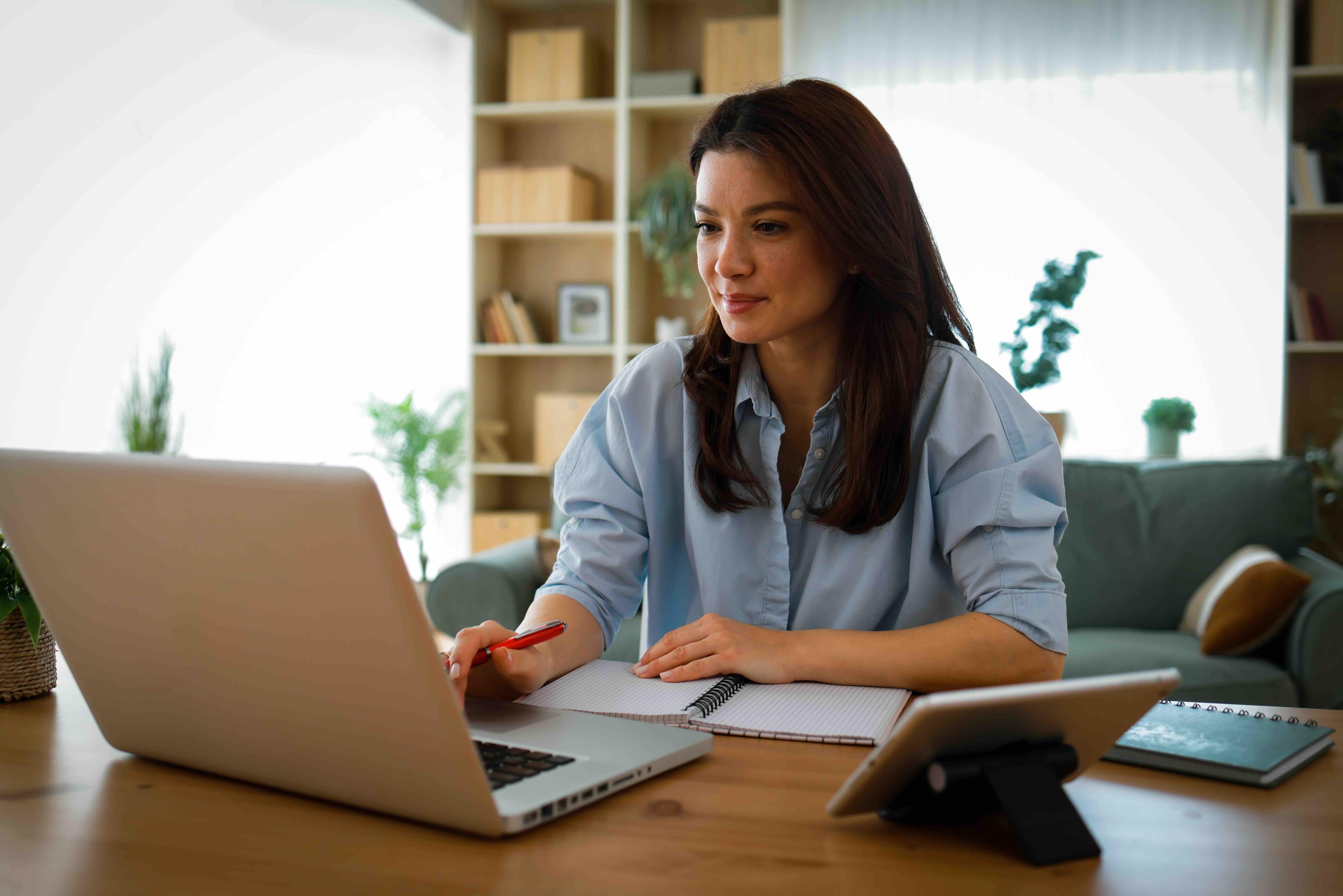 Woman working from home on laptop while taking notes in notebook at wooden desk