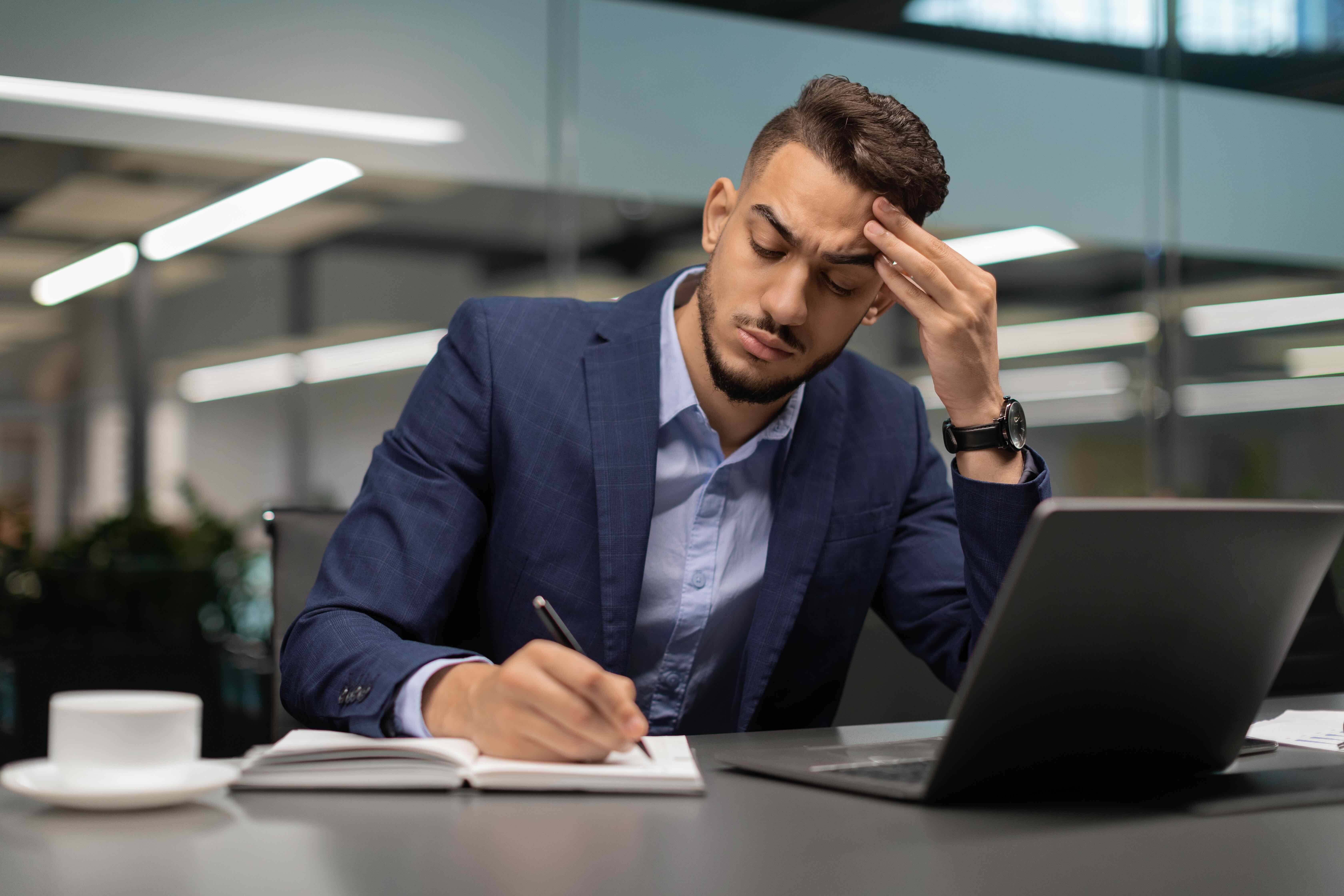 Stressed businessman in suit holding head while writing notes at desk with laptop