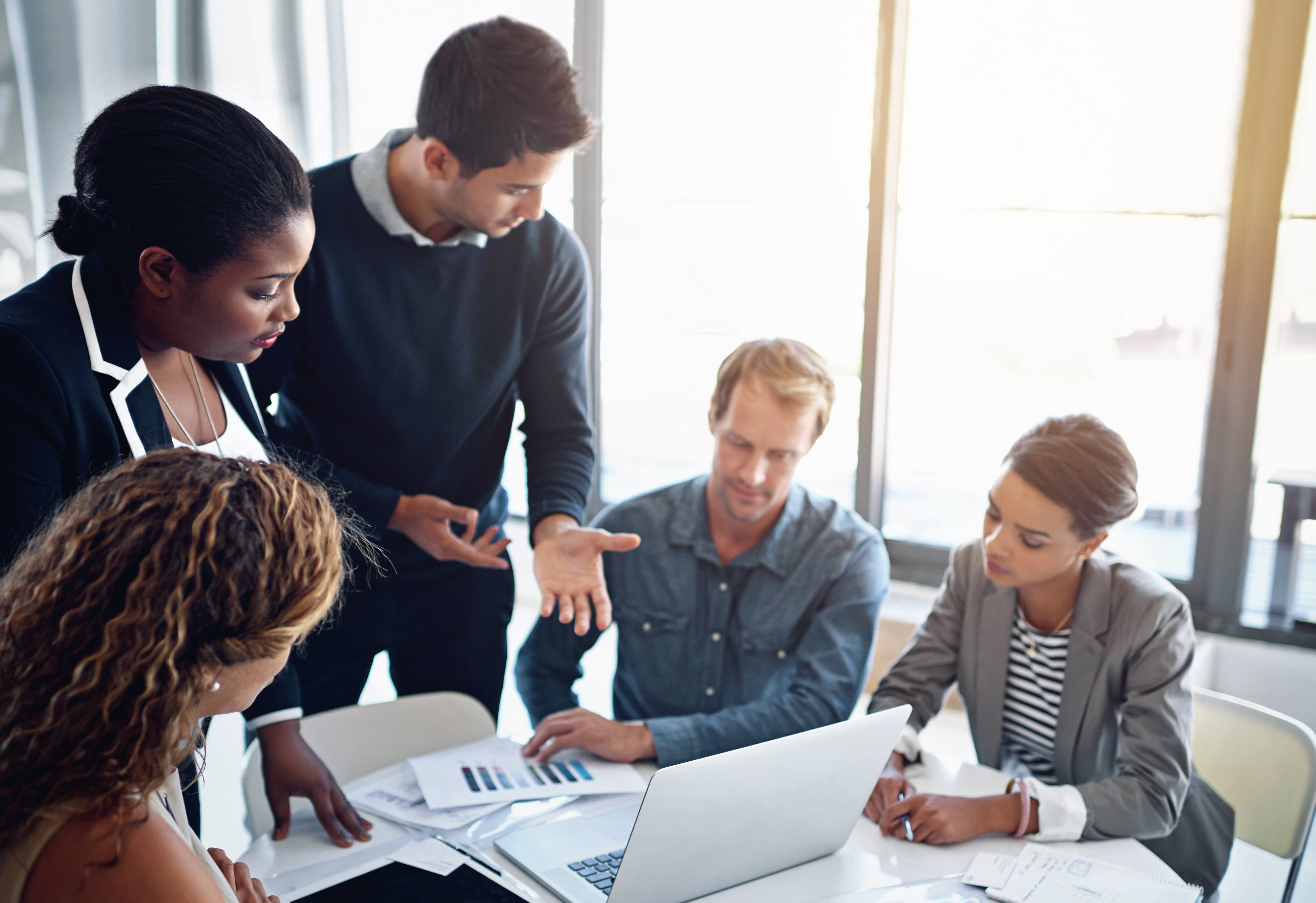 Five diverse business professionals collaborating around a table reviewing charts and documents on a laptop