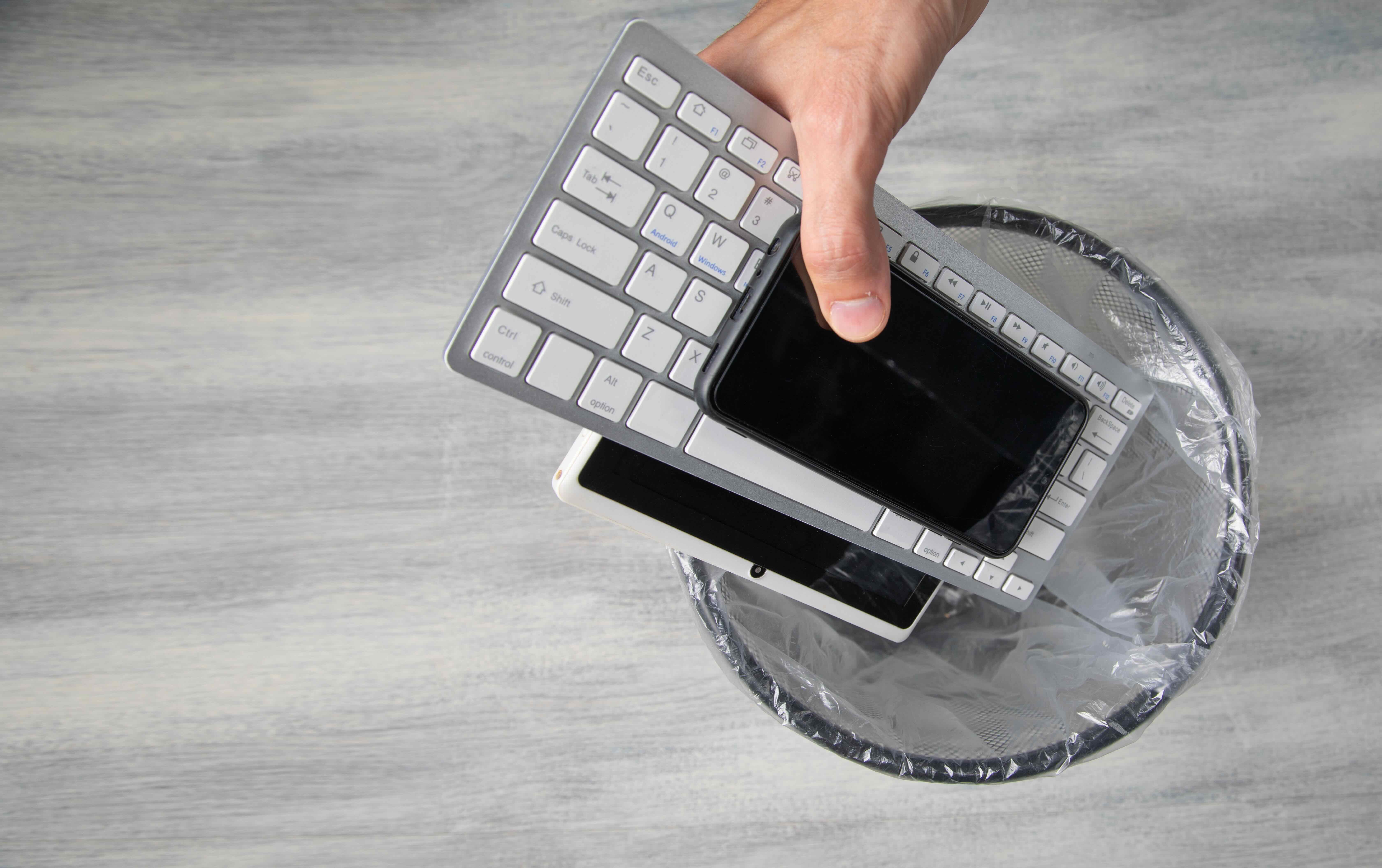 Hand placing smartphone and keyboard into trash bin lined with plastic bag