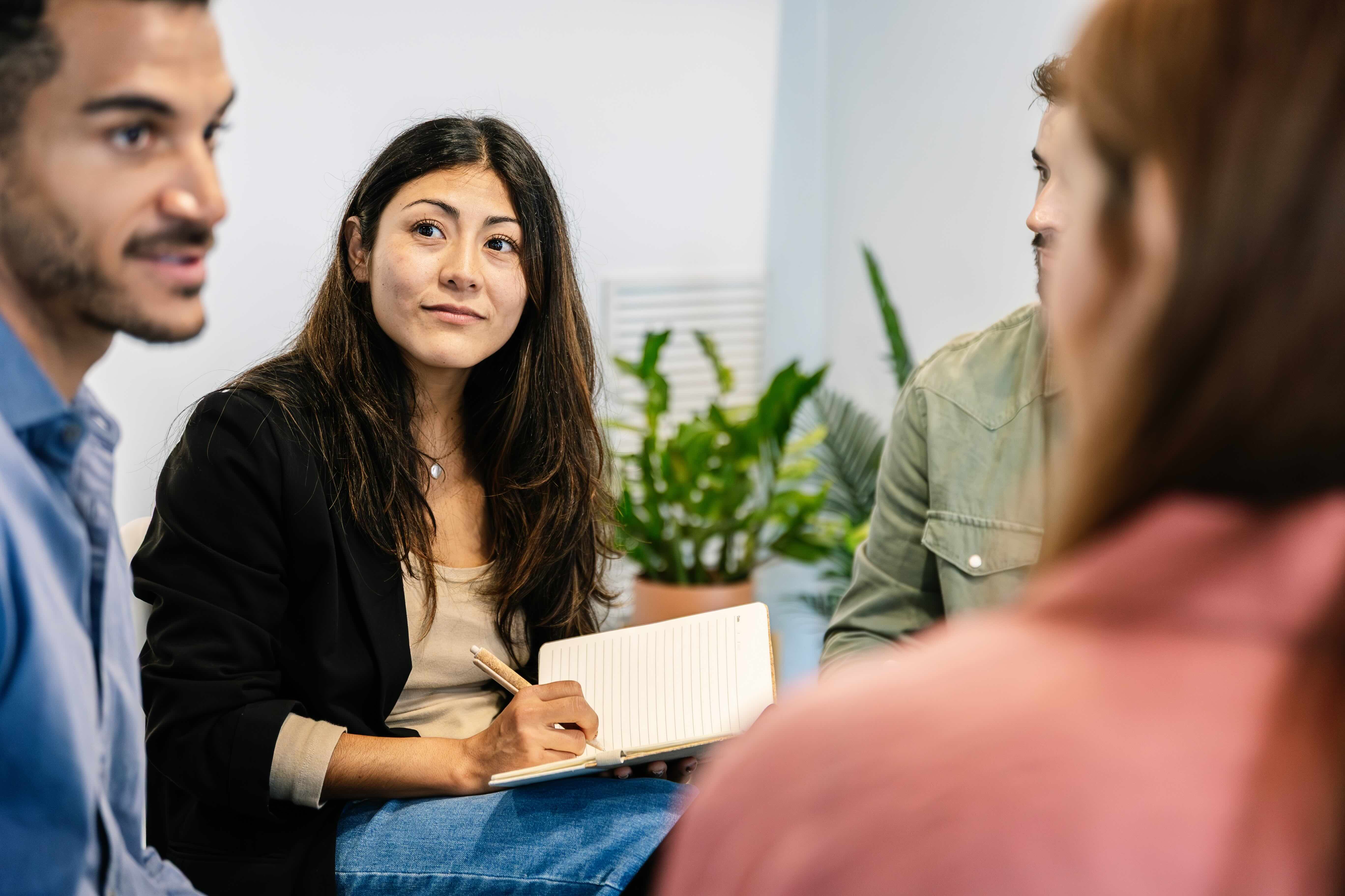 Woman with notebook listens attentively during small group discussion with colleagues in bright office setting