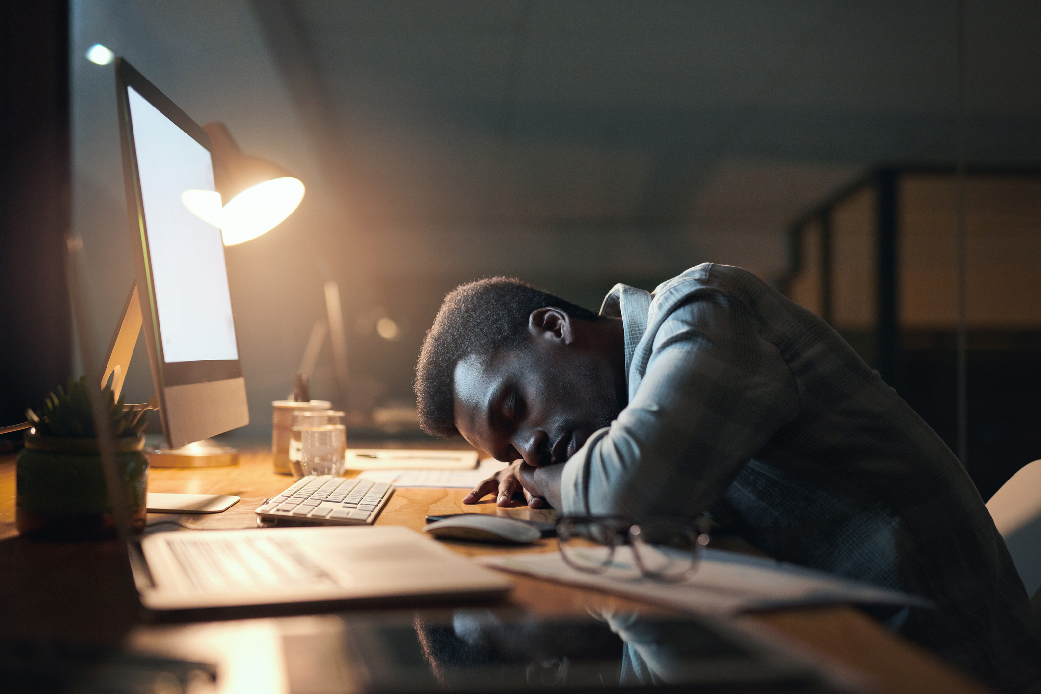 Person exhausted with head down on desk next to computer in dimly lit home office