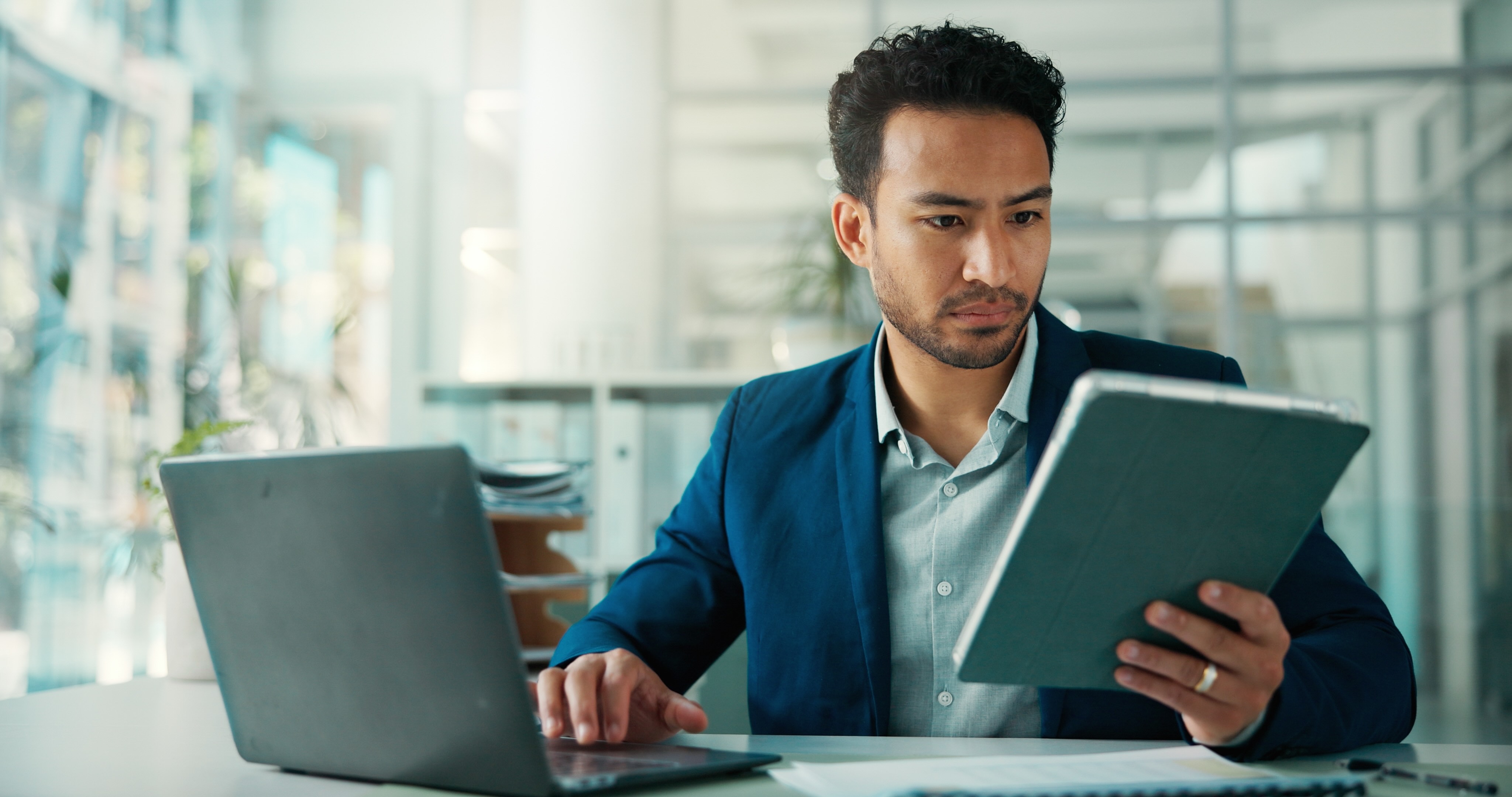 Professional man in blue blazer reviewing tablet while working on laptop in modern office