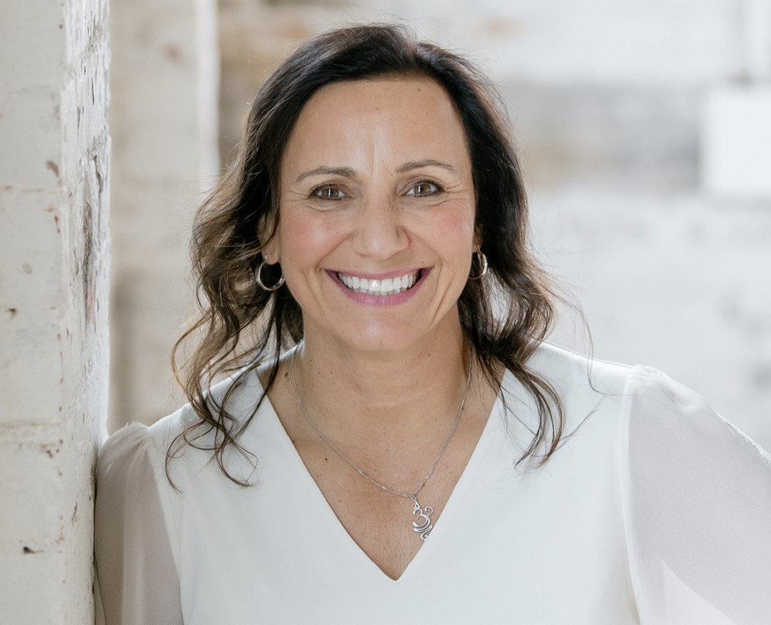 Woman with dark wavy hair wearing white blouse and silver necklace smiling at camera in bright interior setting