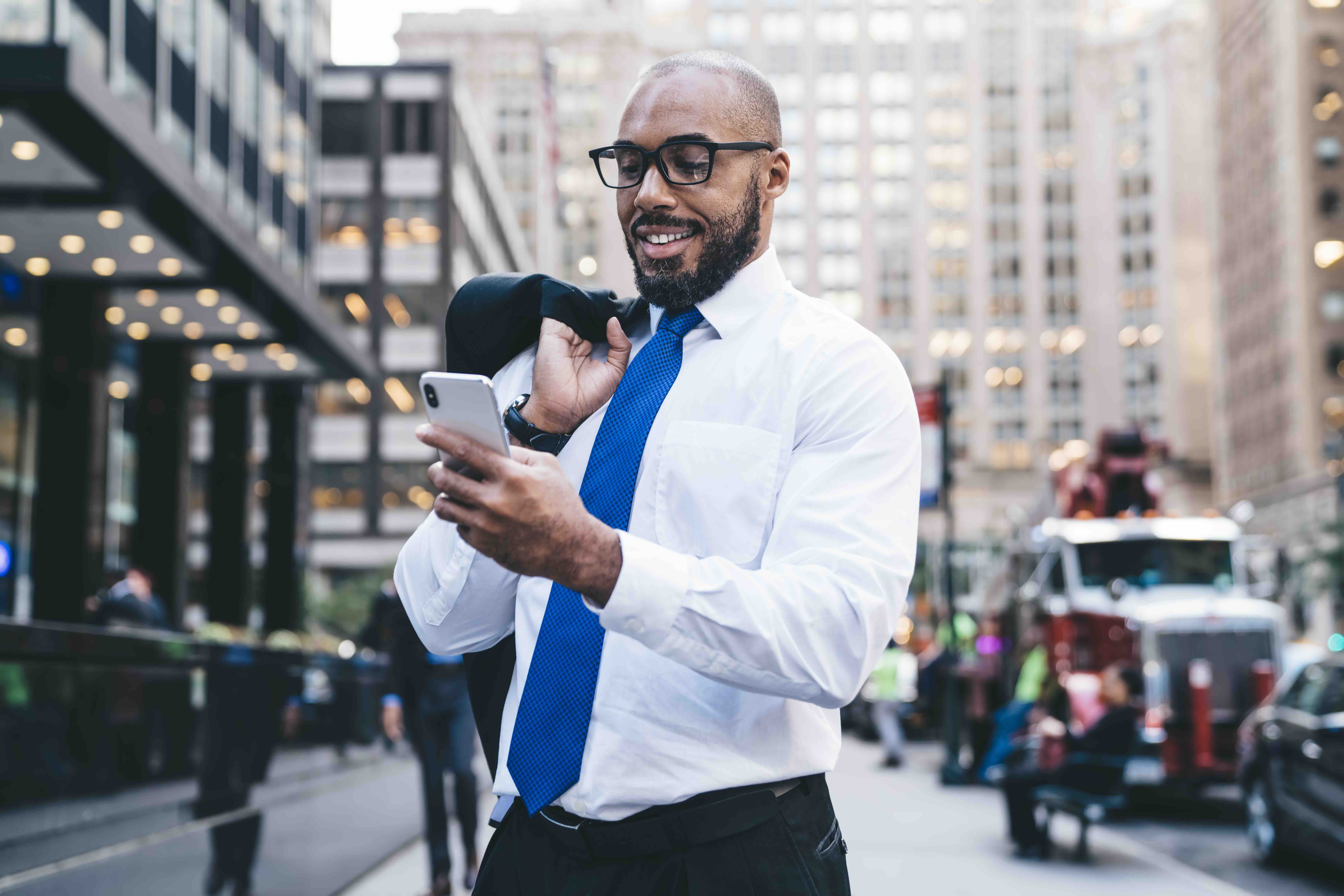 Professional businessman in white shirt and blue tie checks smartphone while walking in urban business district