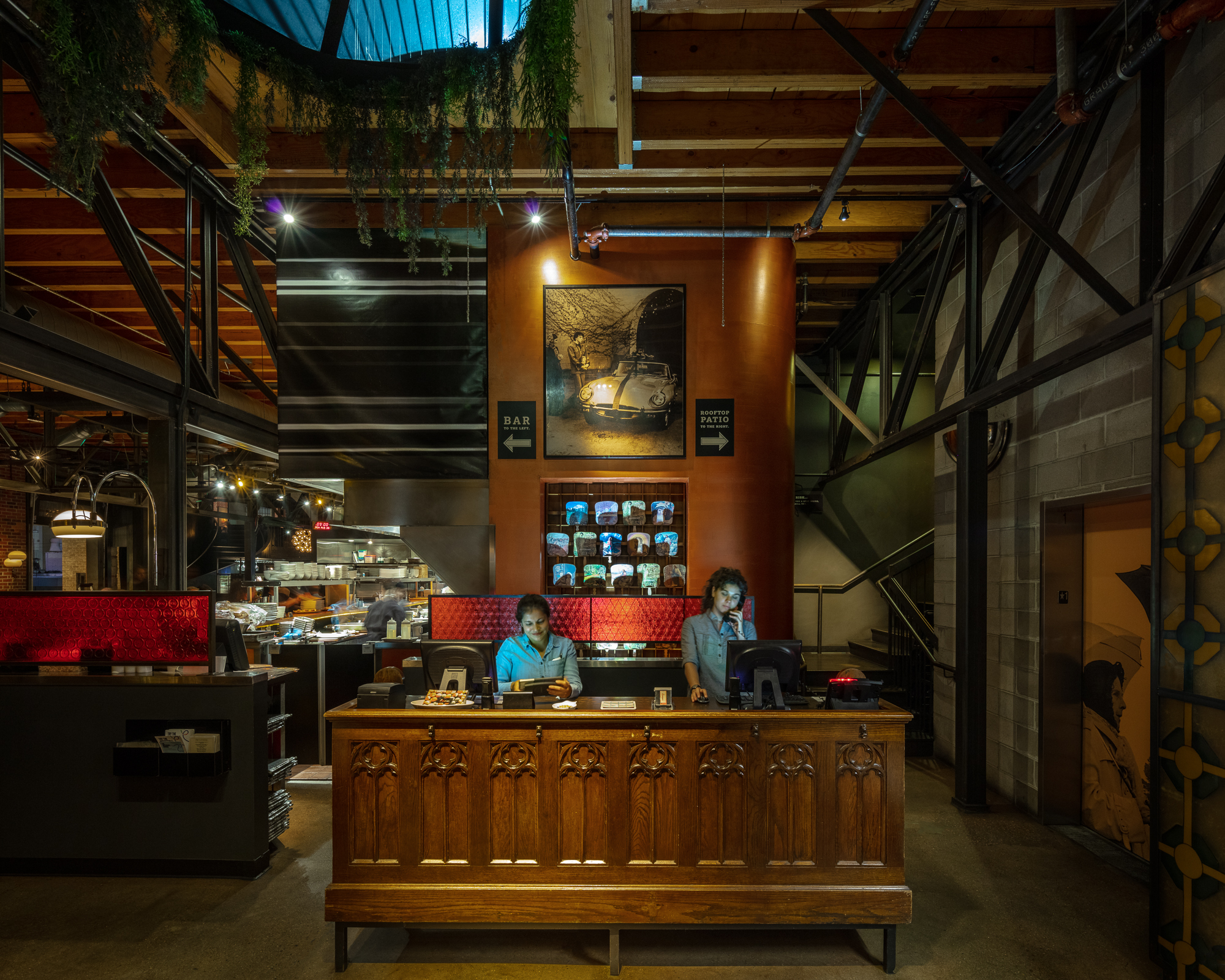 Two staff members work at ornate wooden reception desk in modern industrial restaurant with high ceilings and exposed beams