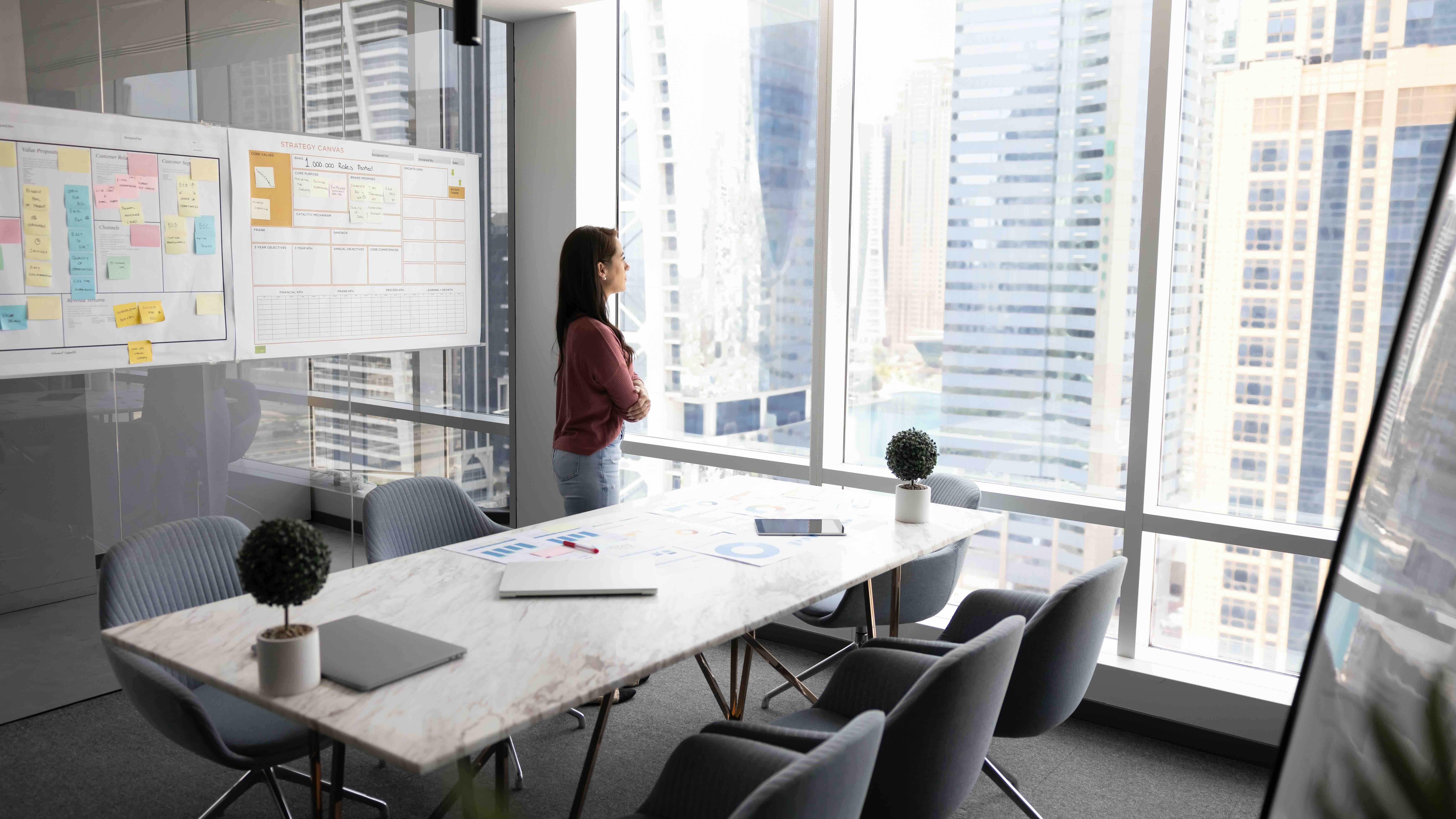 A woman stands alone at a high-rise office window, arms crossed, beside an empty conference table and strategy boards