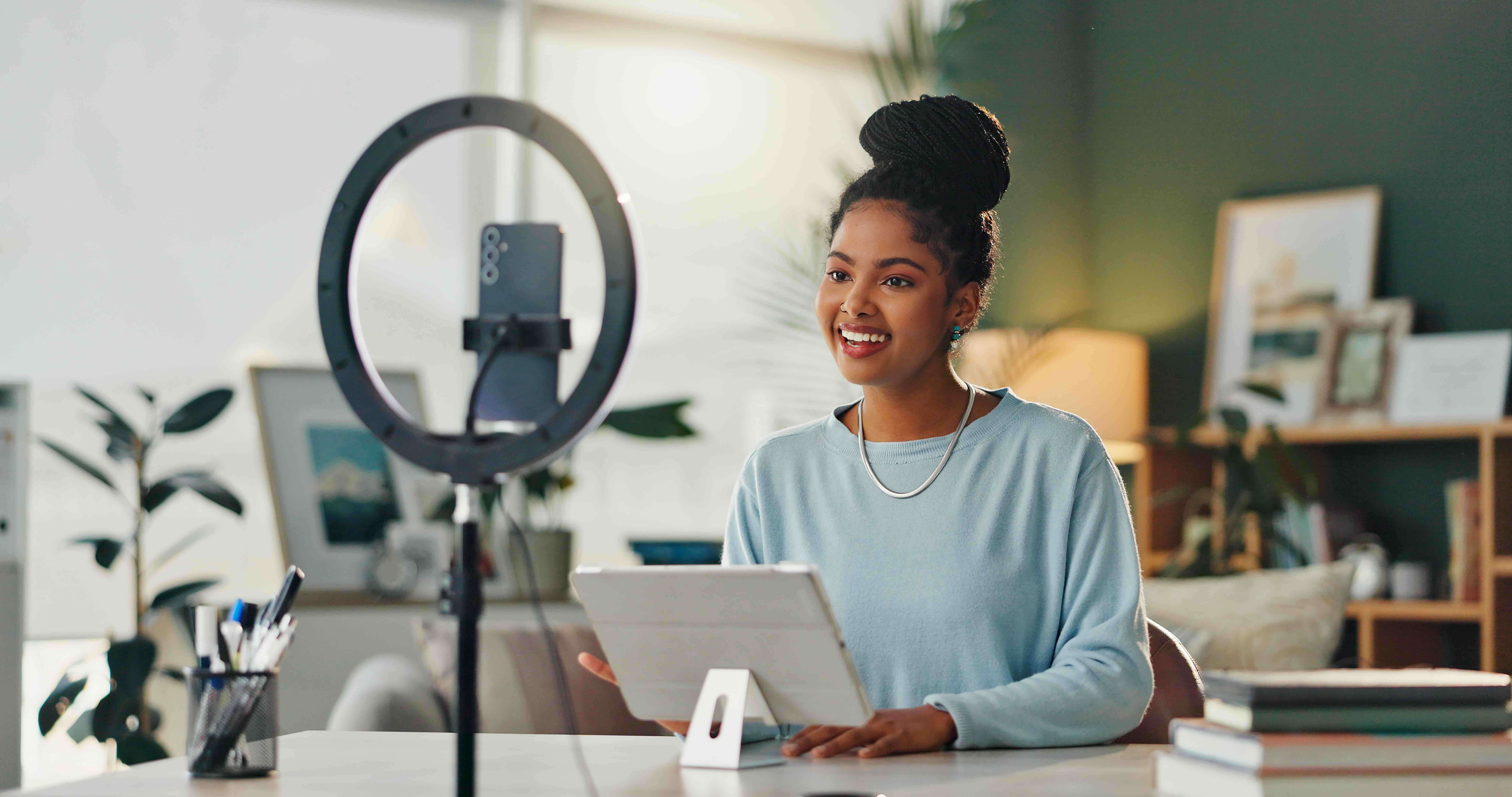 A woman films content with a smartphone and ring light while smiling and holding a tablet at her desk.