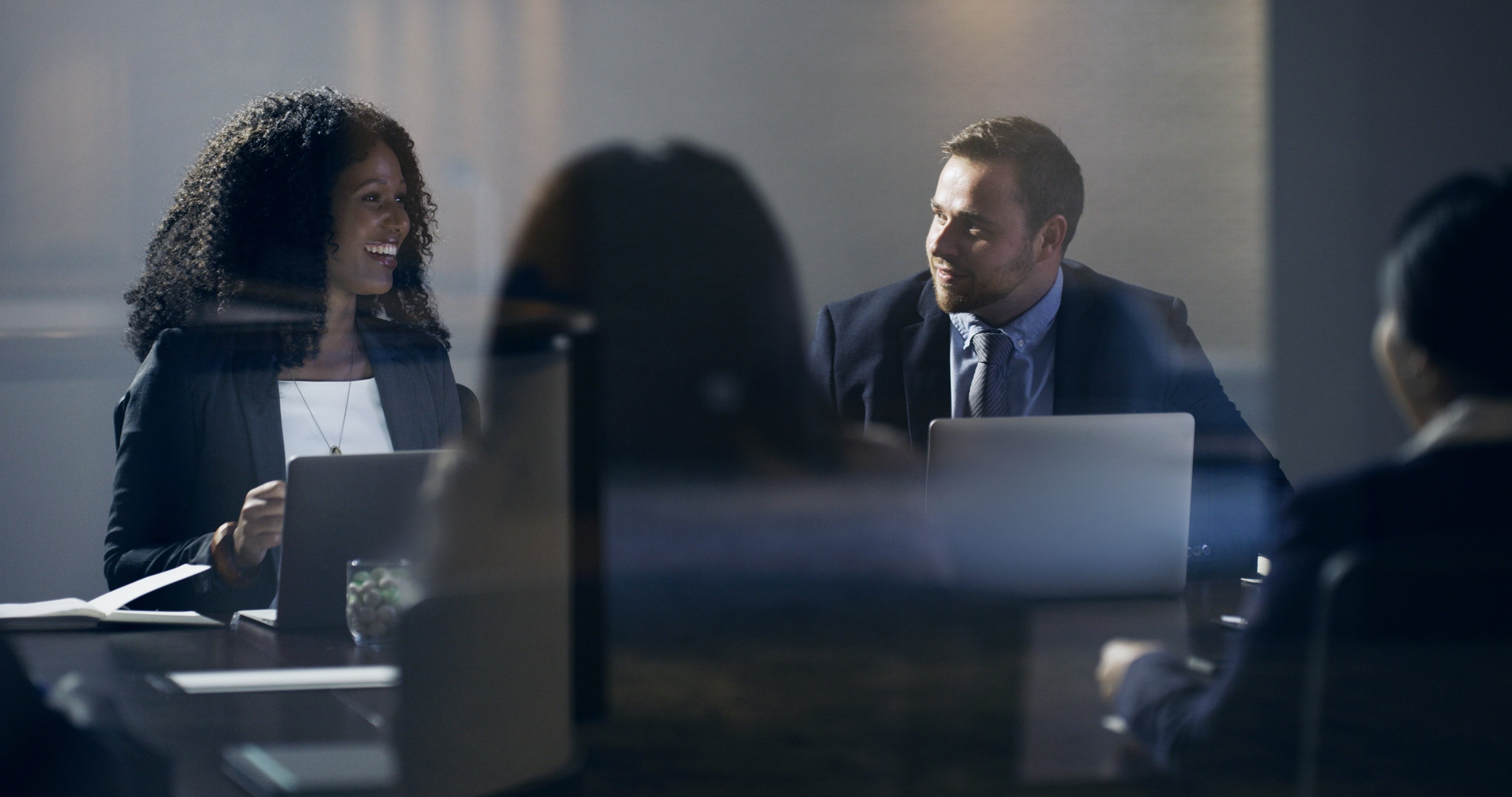 Two professionals smiling and engaged in collaborative discussion during a business meeting with laptops open