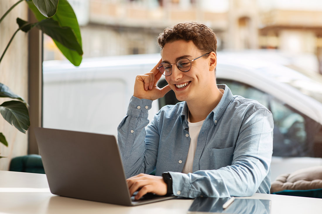 young man with curly hair and glasses smiling at his laptop in a bright room with a plant