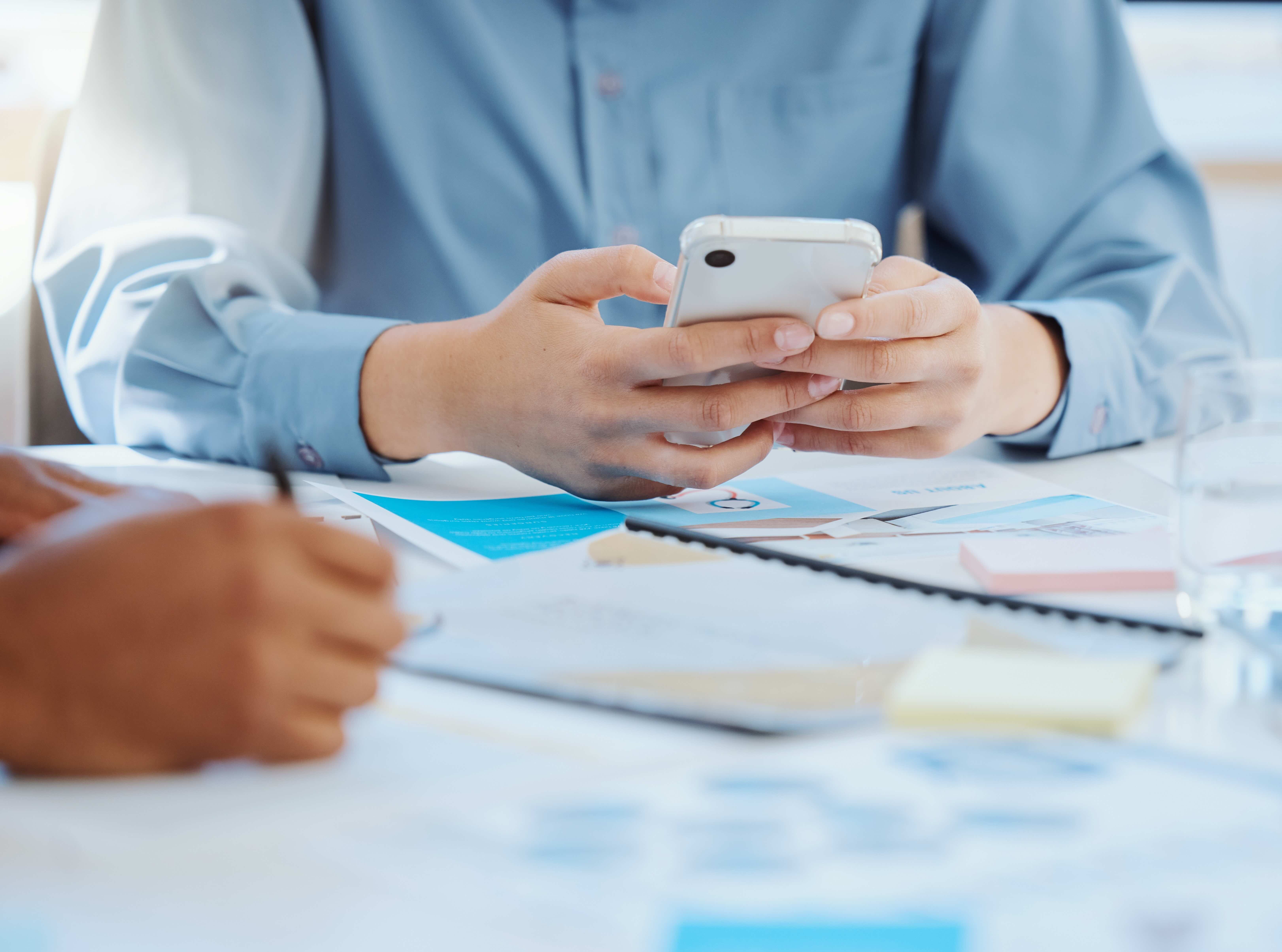 Person in light blue shirt holding white smartphone during business meeting with documents spread on table