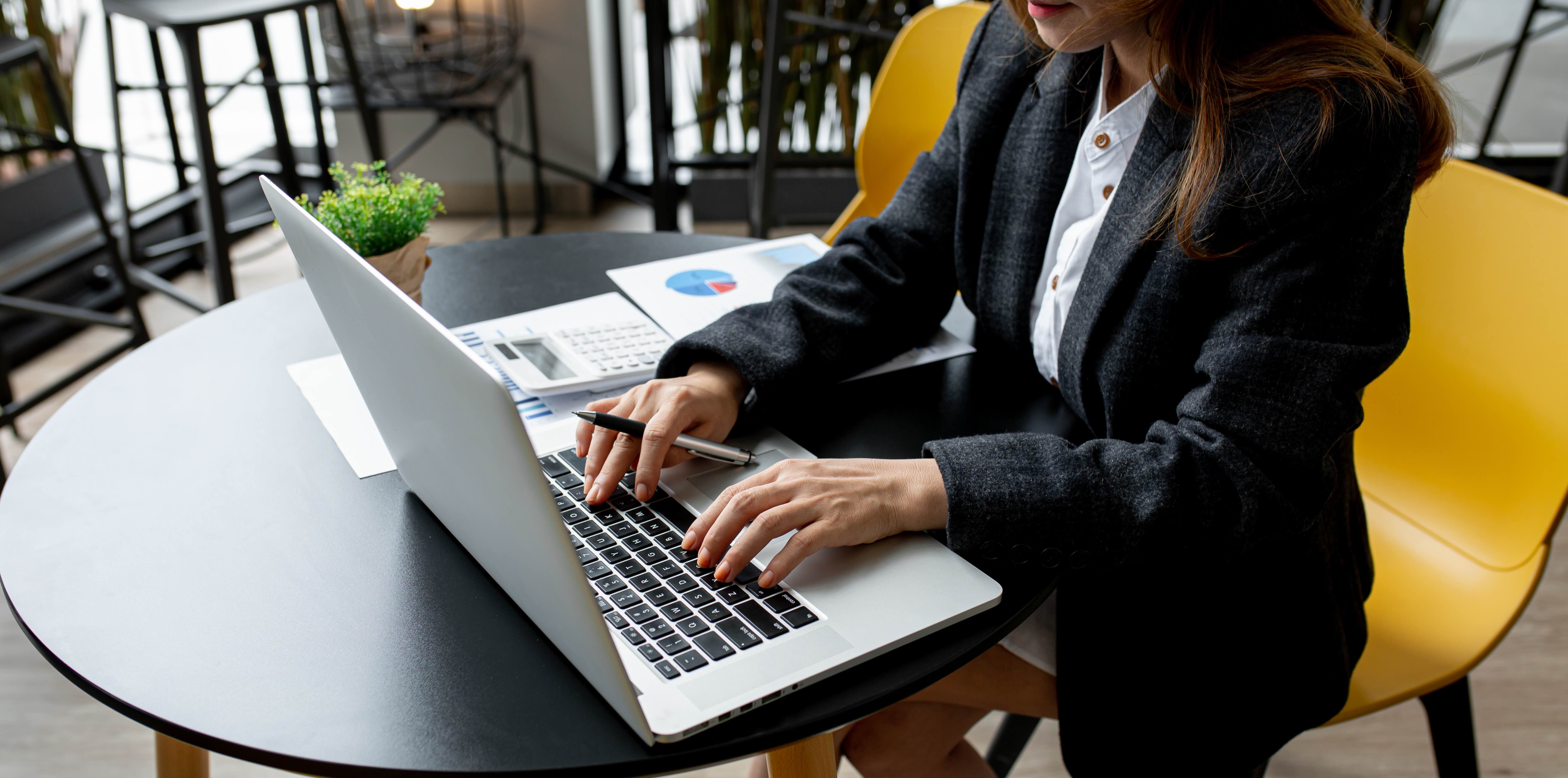 Professional woman in dark blazer typing on laptop at modern workspace with financial documents and pie chart visible