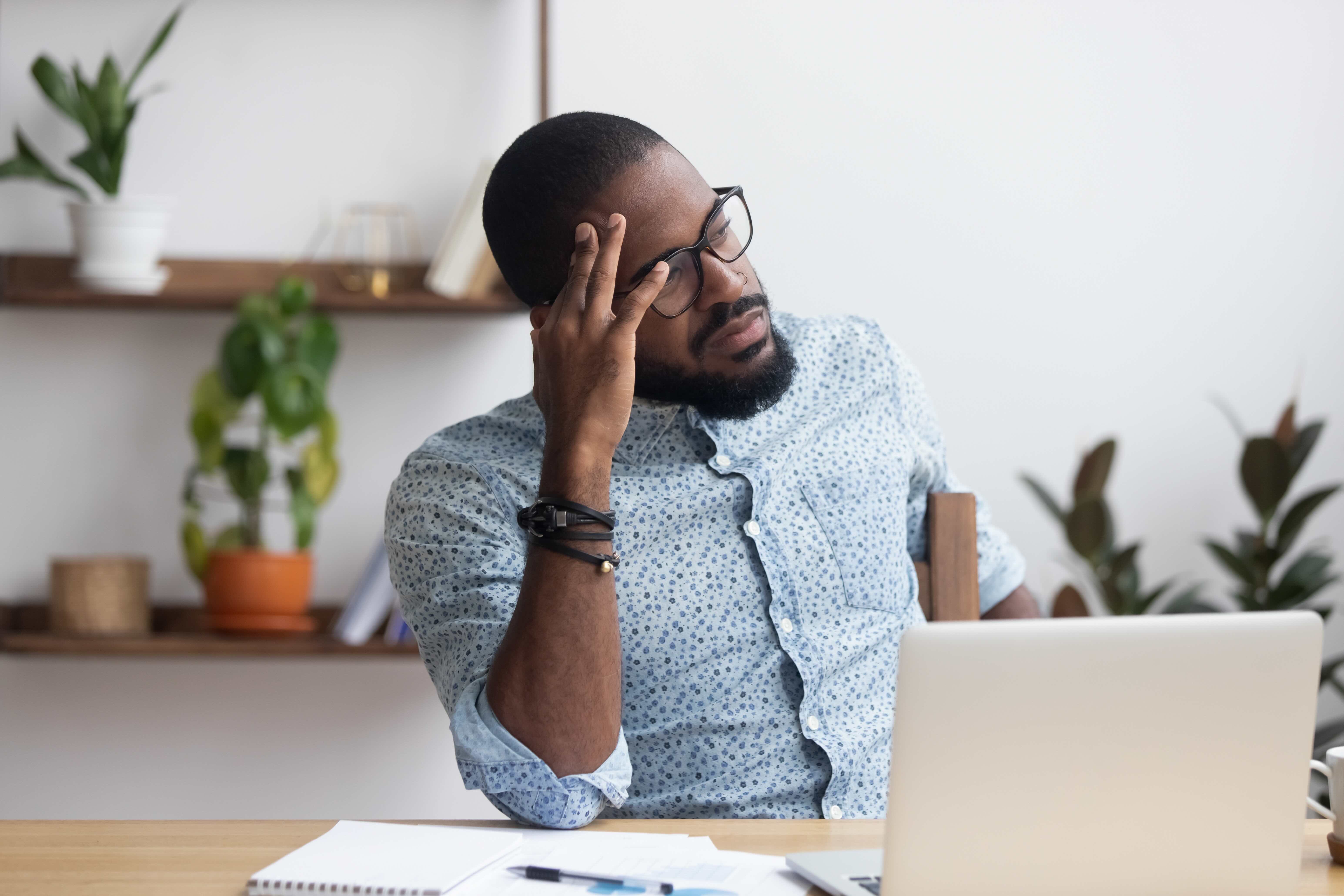 Exhausted professional with glasses rubbing temple while sitting at desk, showing signs of burnout and emotional fatigue