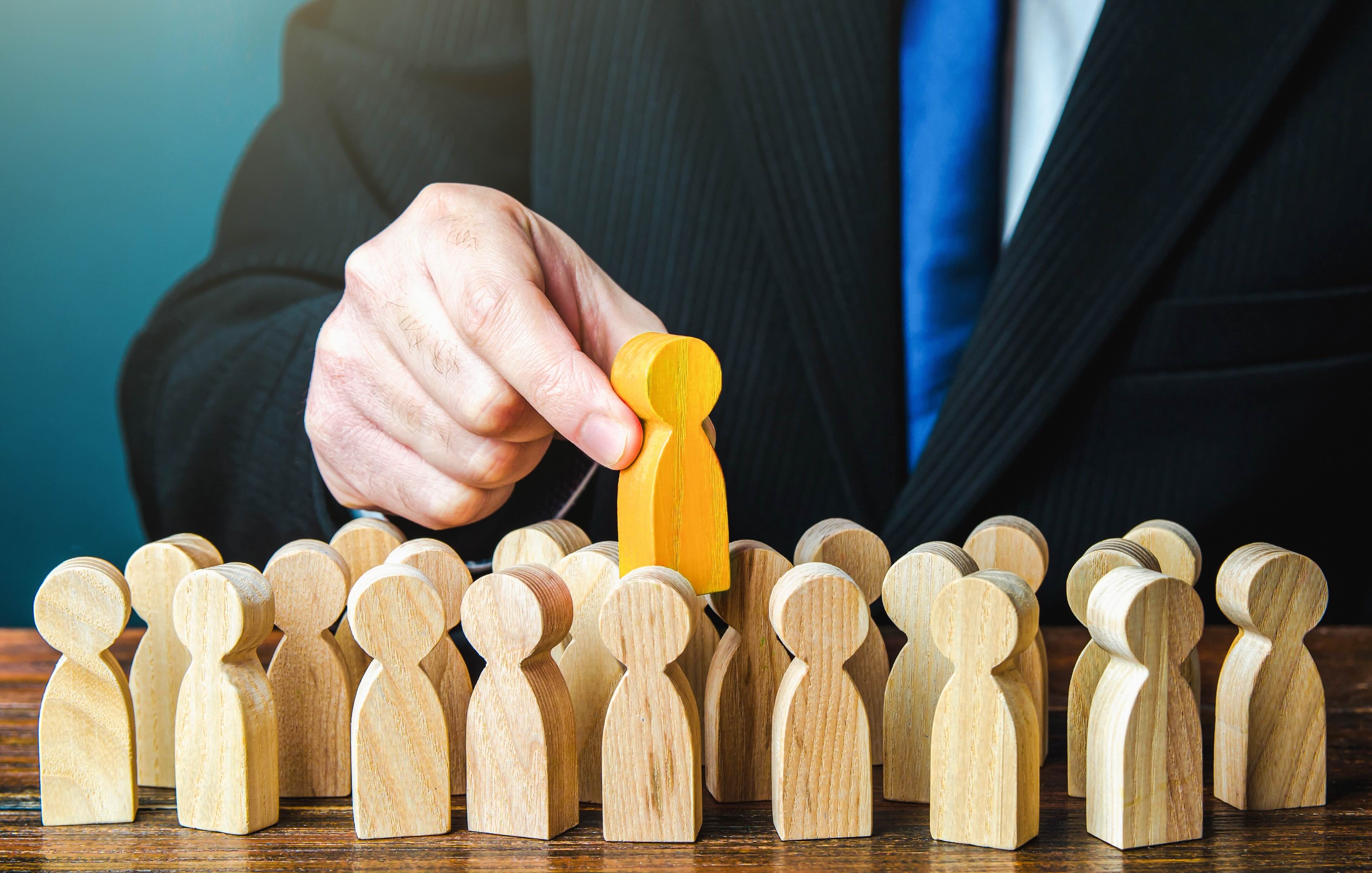 Businessperson selecting a yellow wooden figure from a crowd of natural wooden figures representing recruitment and hiring