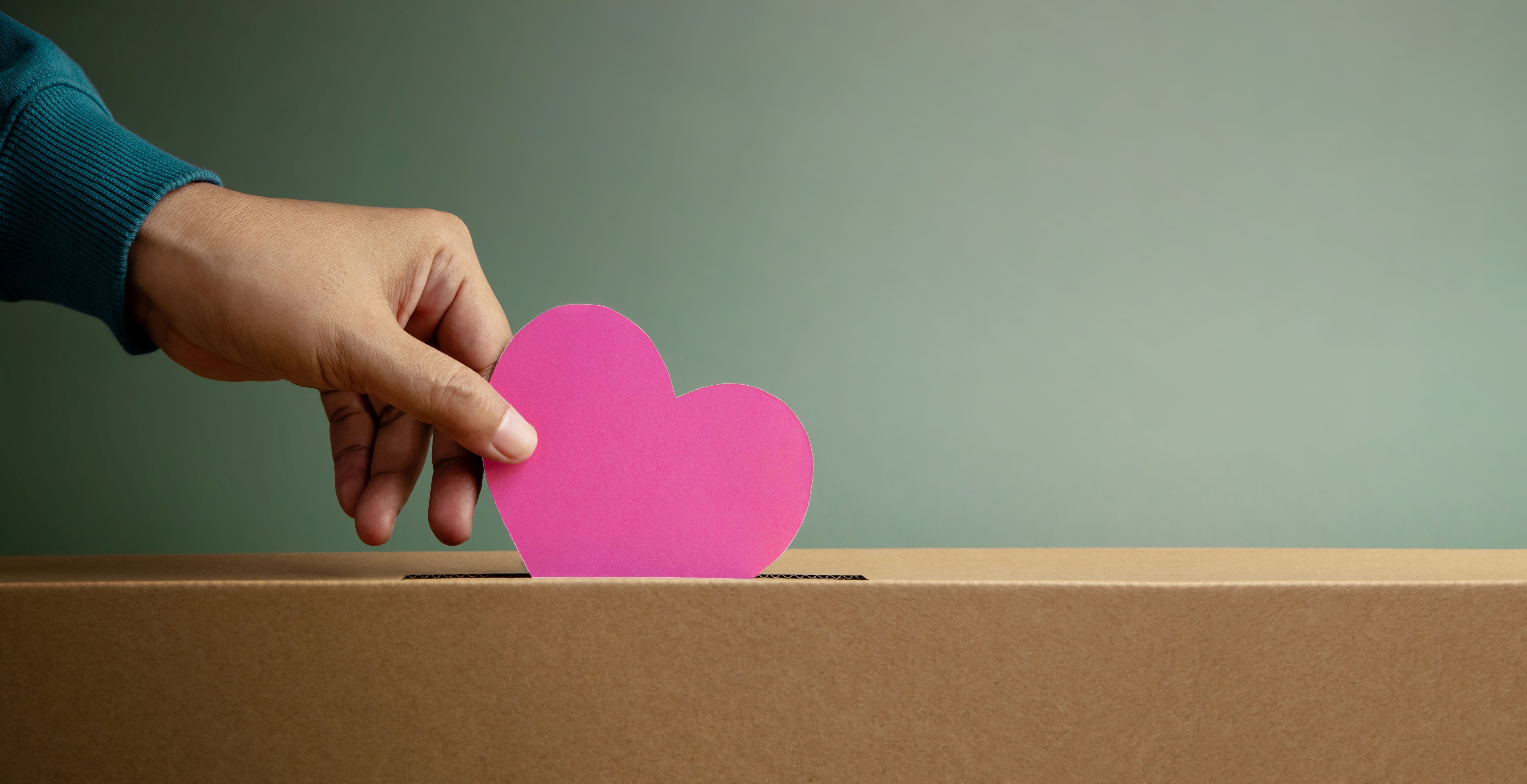 Hand placing pink heart-shaped paper into cardboard donation box against neutral background