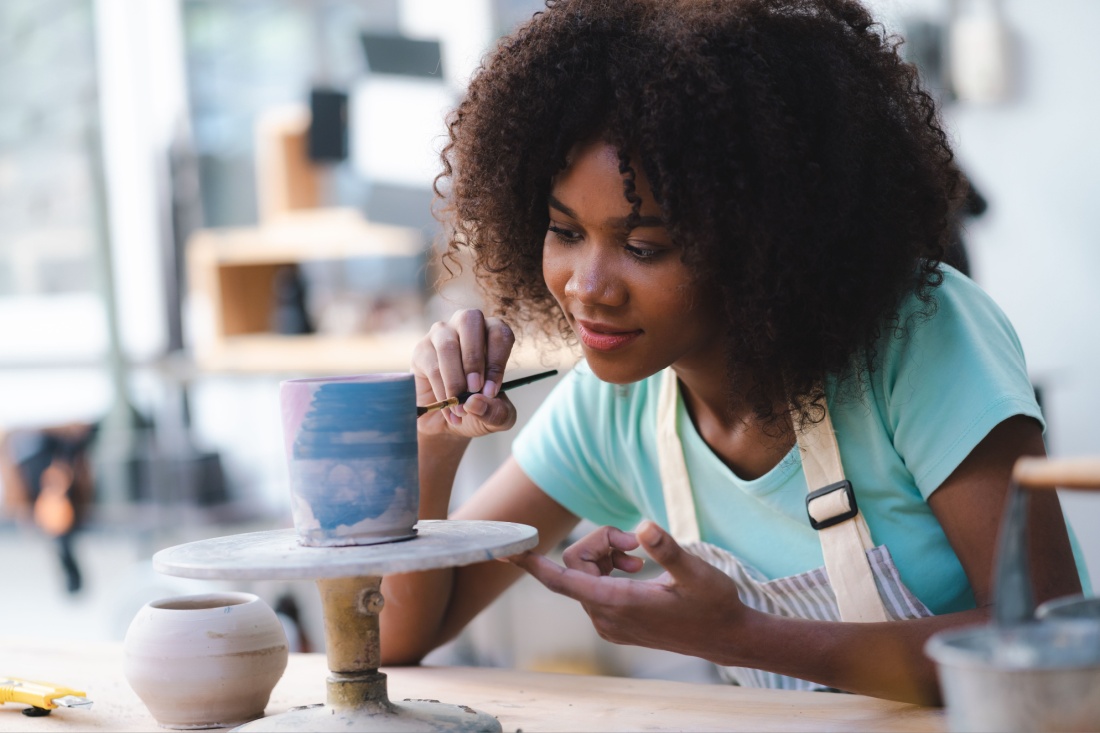 Woman in teal shirt and apron carefully painting pottery at a workshop table