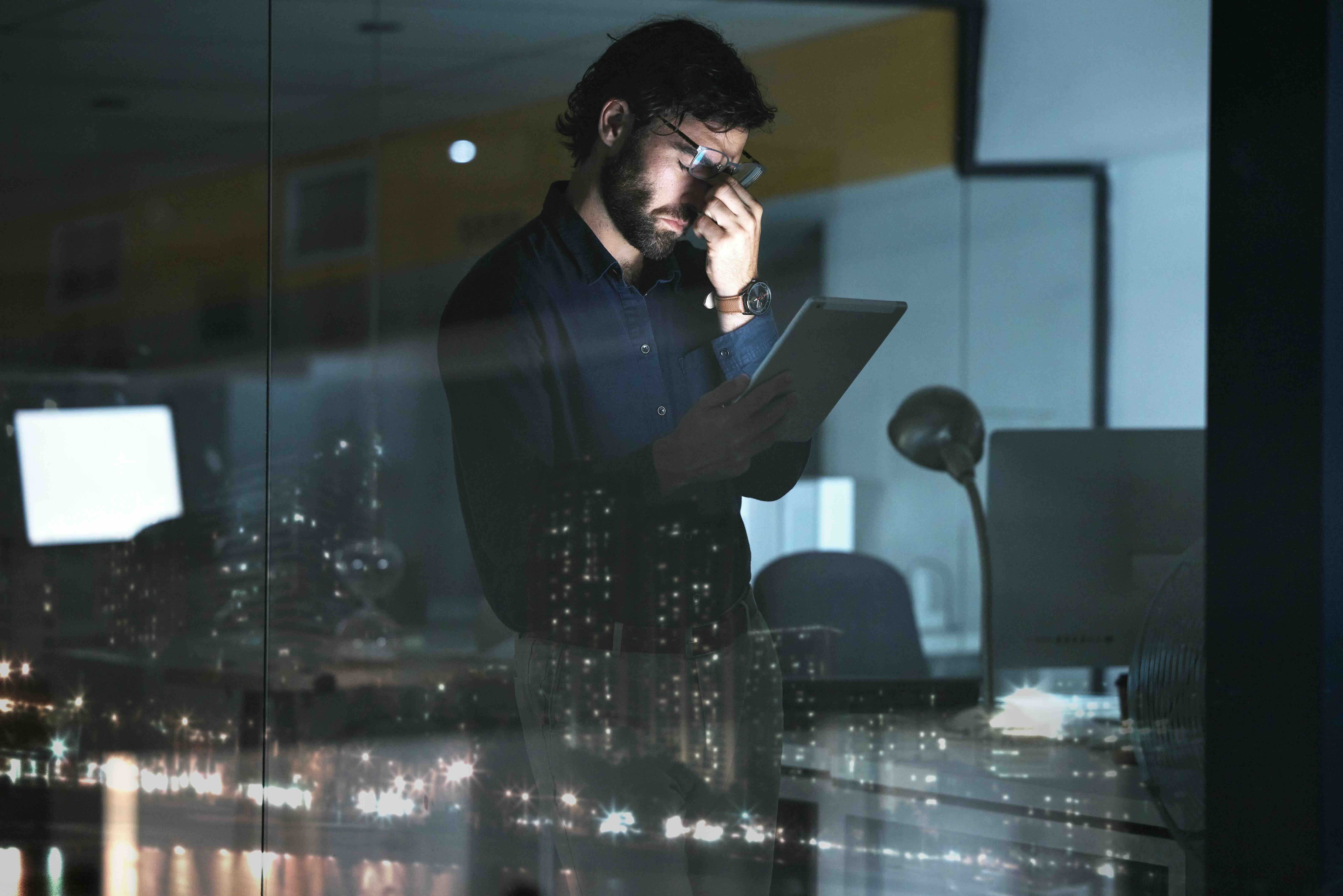 Stressed executive with glasses looking down at tablet in dimly lit office with city lights reflected in window