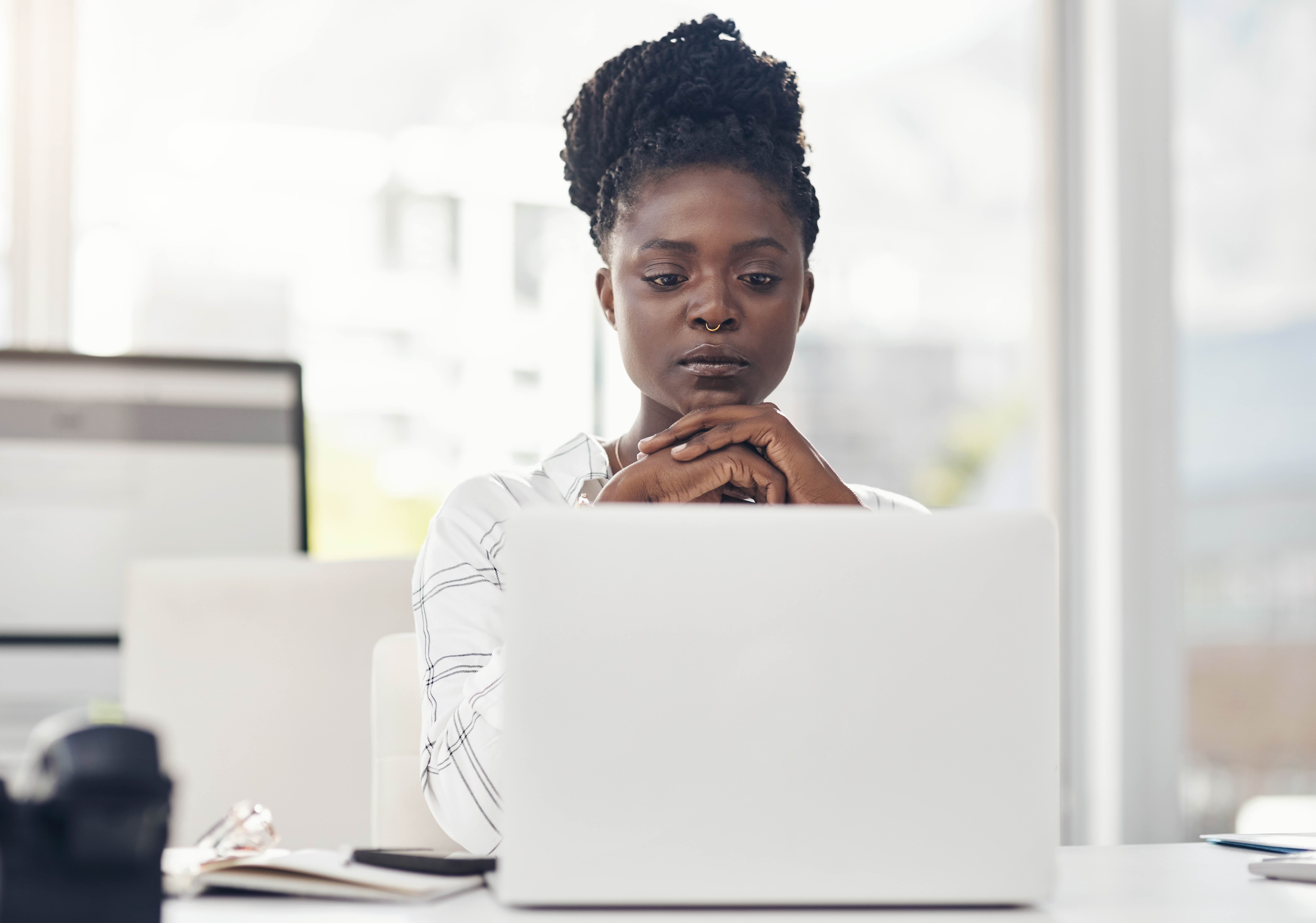 Woman in white shirt looking pensively at laptop screen while resting chin on hand at office desk