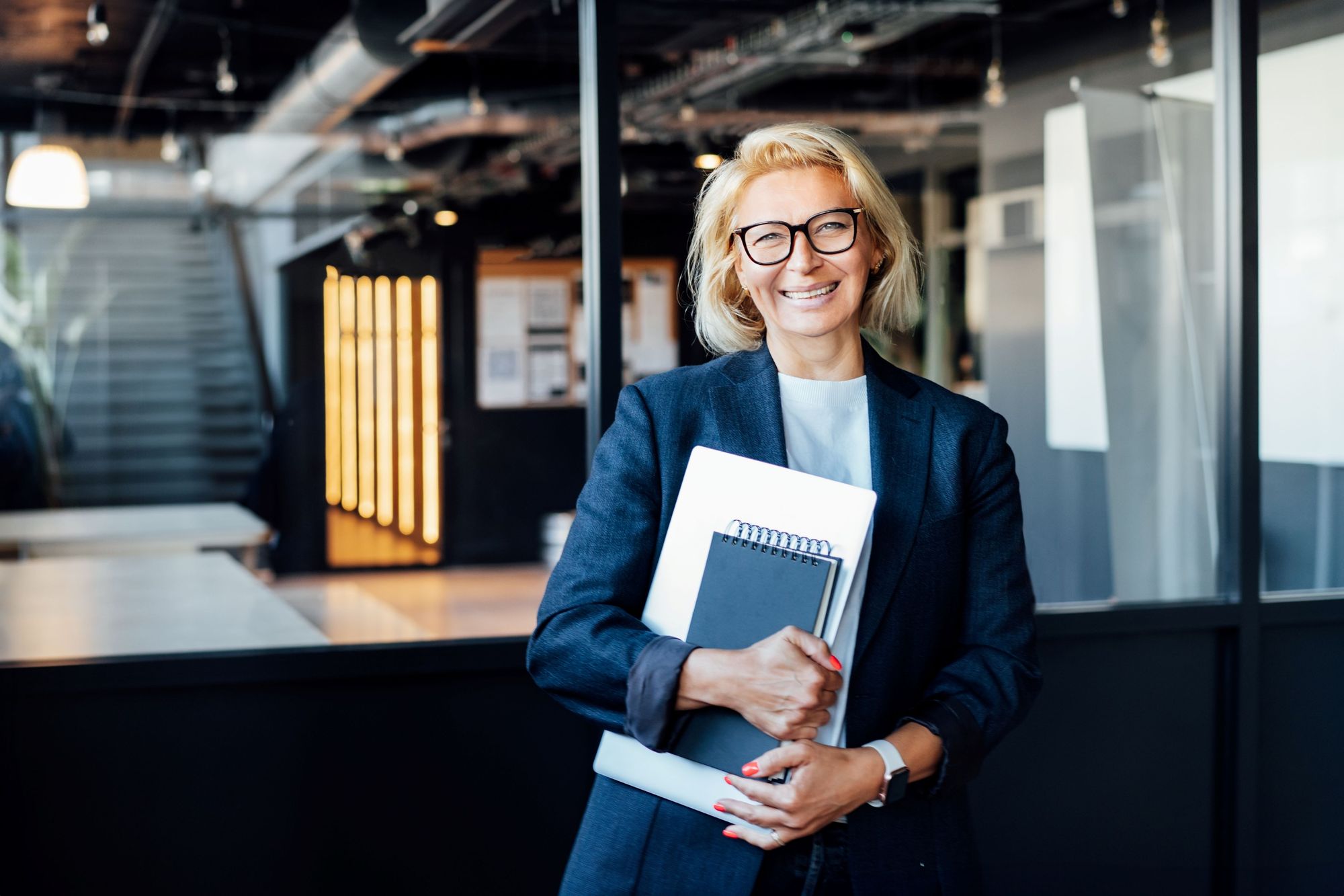 Professional woman in navy blazer holding notebooks and smiling confidently in modern office setting