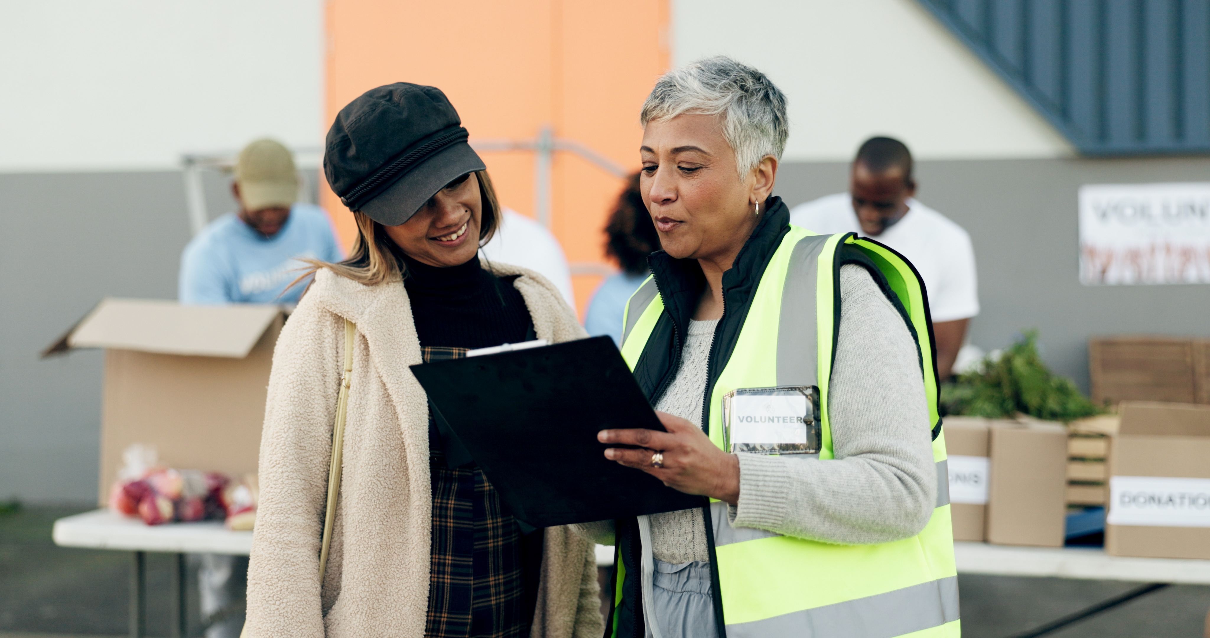 Two women reviewing documents on clipboard in volunteer warehouse with donation boxes and team members working in background