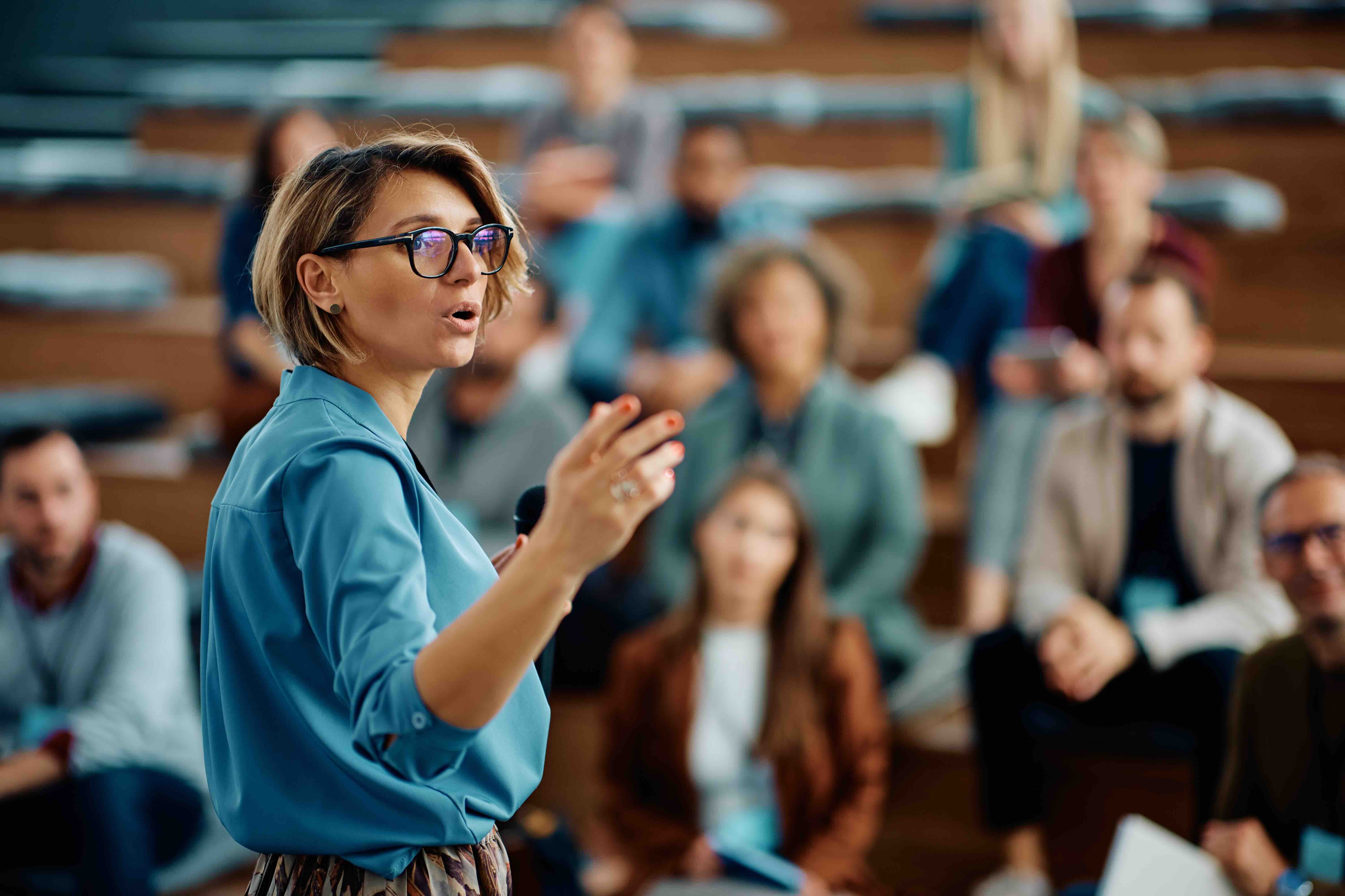 Mid adult businesswoman confidently talking to group of seminar attendees in conference hall.