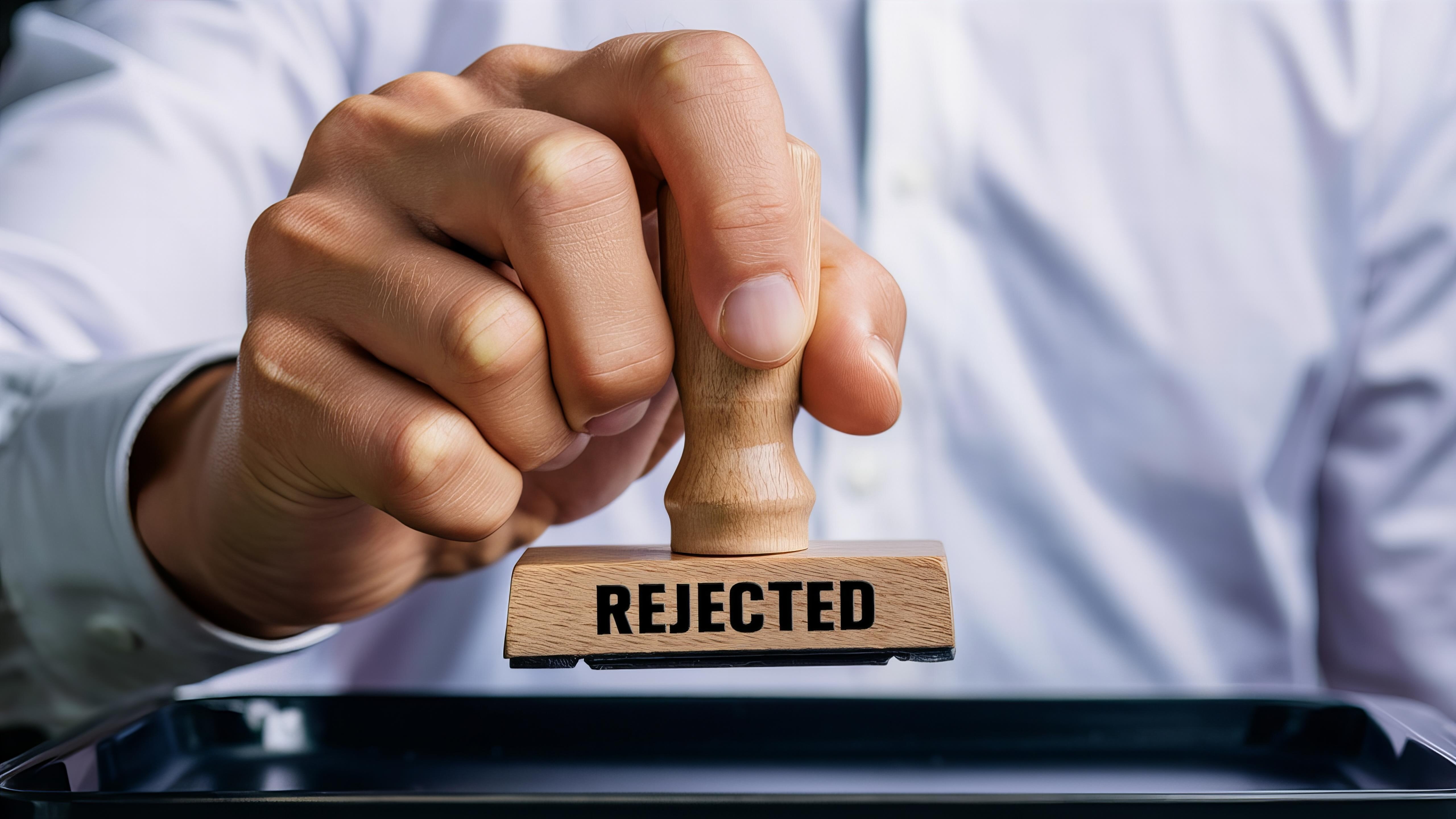 Hand holding wooden stamp with "REJECTED" text over document on desk