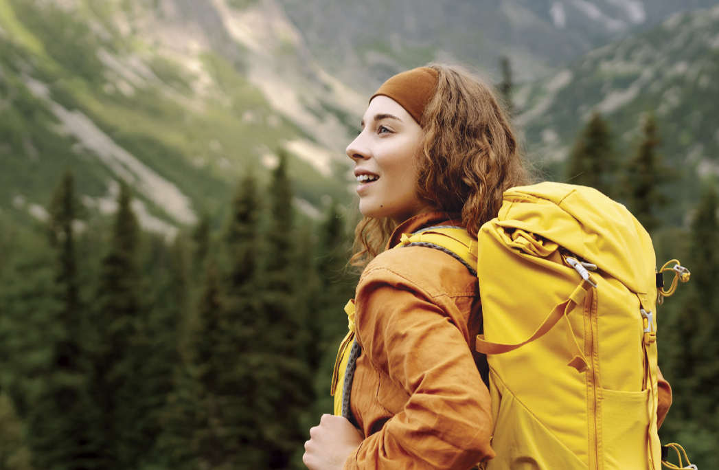 A woman in an orange jacket with a yellow hiking backpack gazes out over a forested mountain landscape