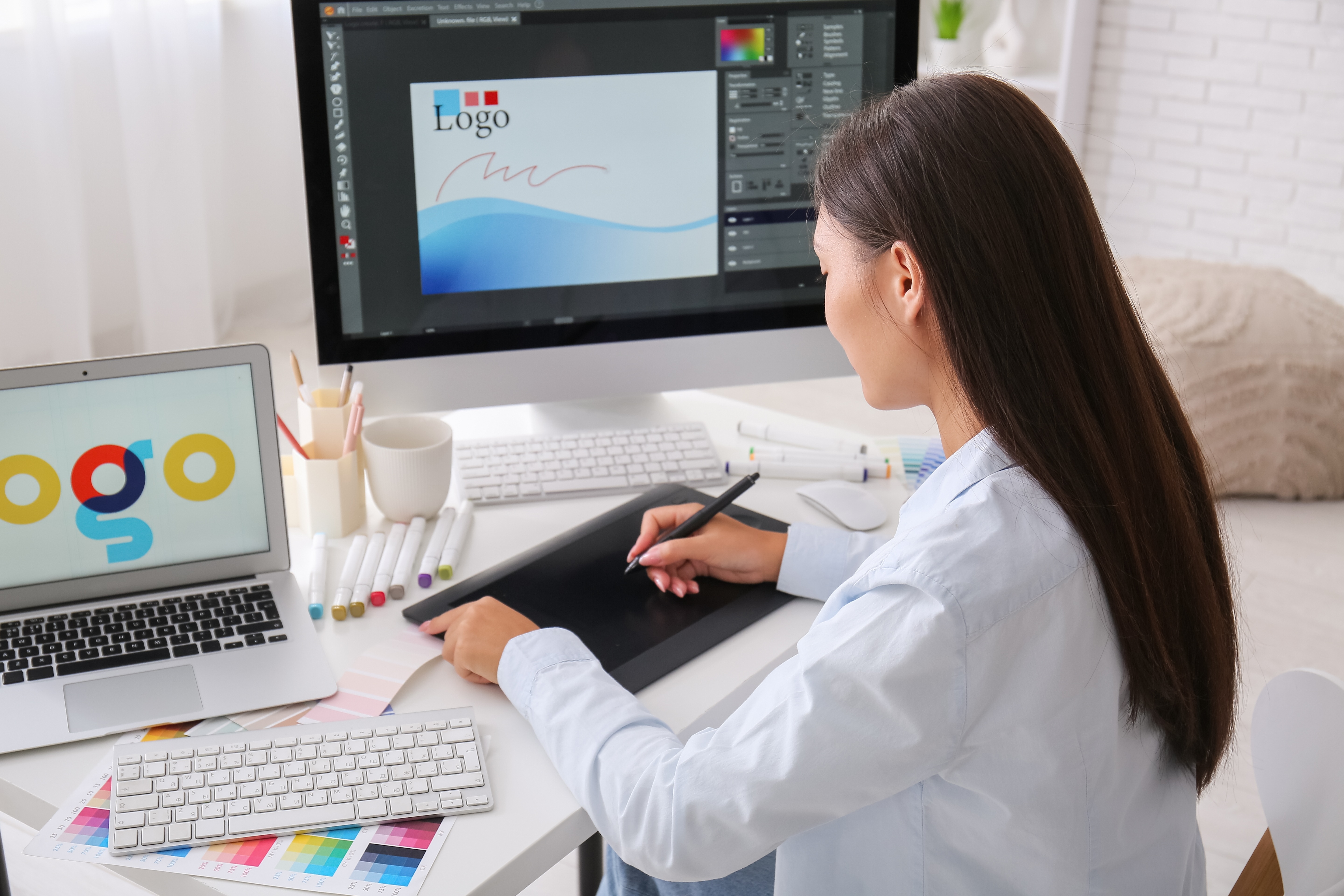 Graphic designer using digital tablet to create logo at desk with laptop and color swatches
