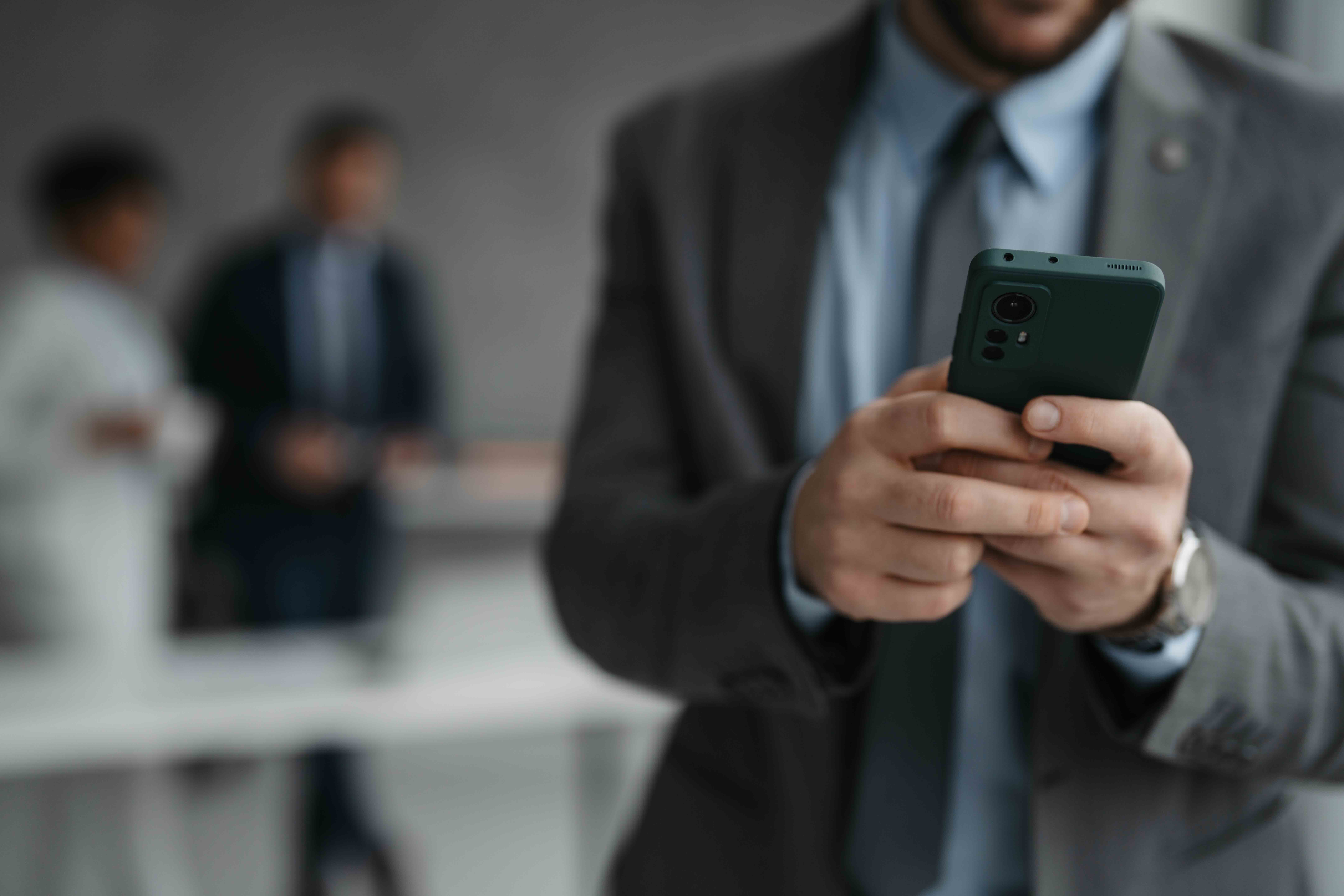 4:12 PMClaude responded: A man in a dark suit and tie holds a dark green smartphone in both hands in the foreground, with two blurred figures standing in a gray office space behind him.A man in a dark suit and tie holds a dark green smartphone in both hands in the foreground, with two blurred figures standing in a gray office space behind him.