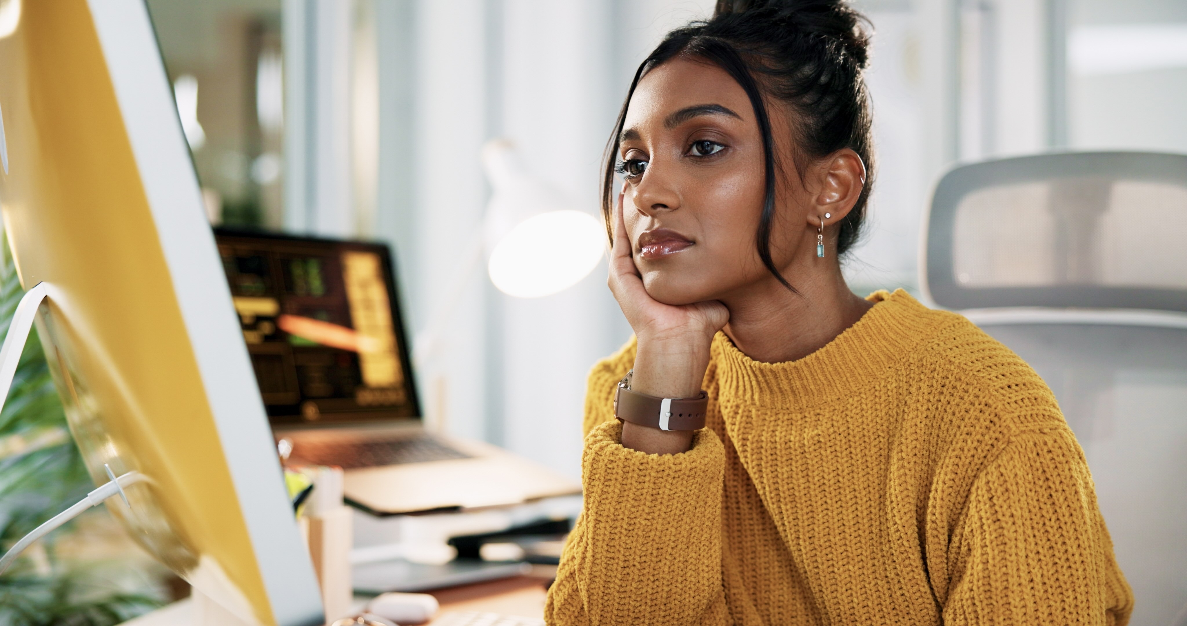 Woman in yellow sweater looking thoughtfully at computer screen while resting chin on hand