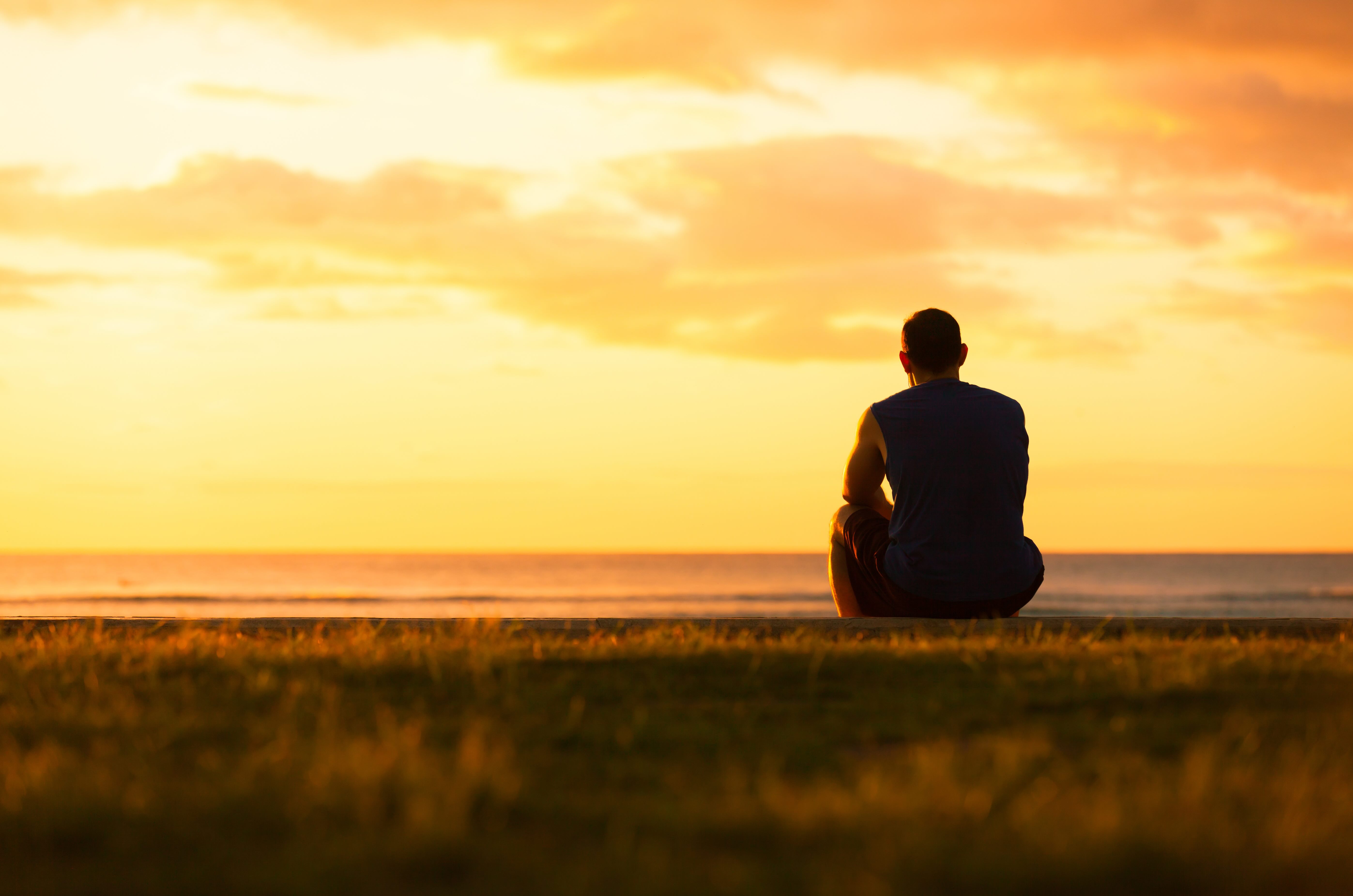 Person sitting alone on grass overlooking ocean at sunset, contemplating in solitude