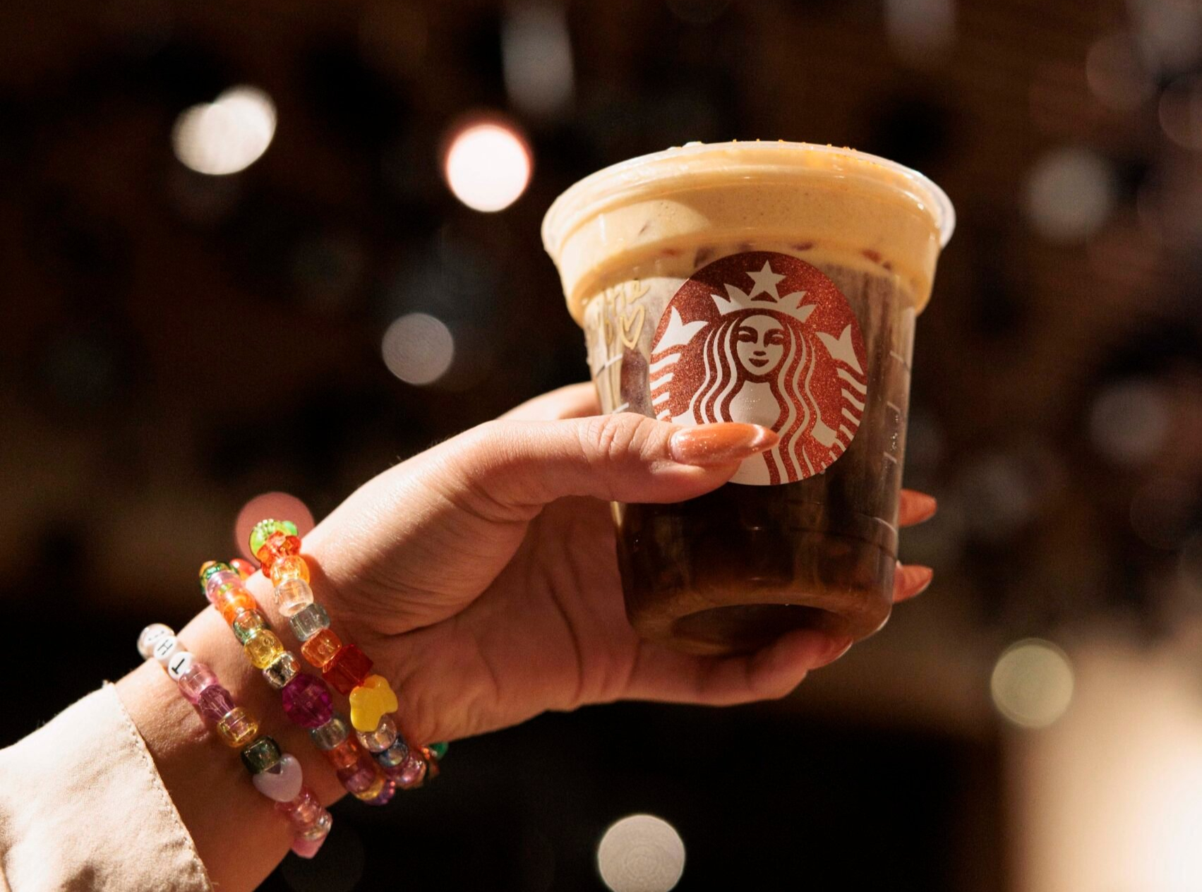 Hand holding a Starbucks cold brew with foam topping and colorful beaded bracelets against a blurred background