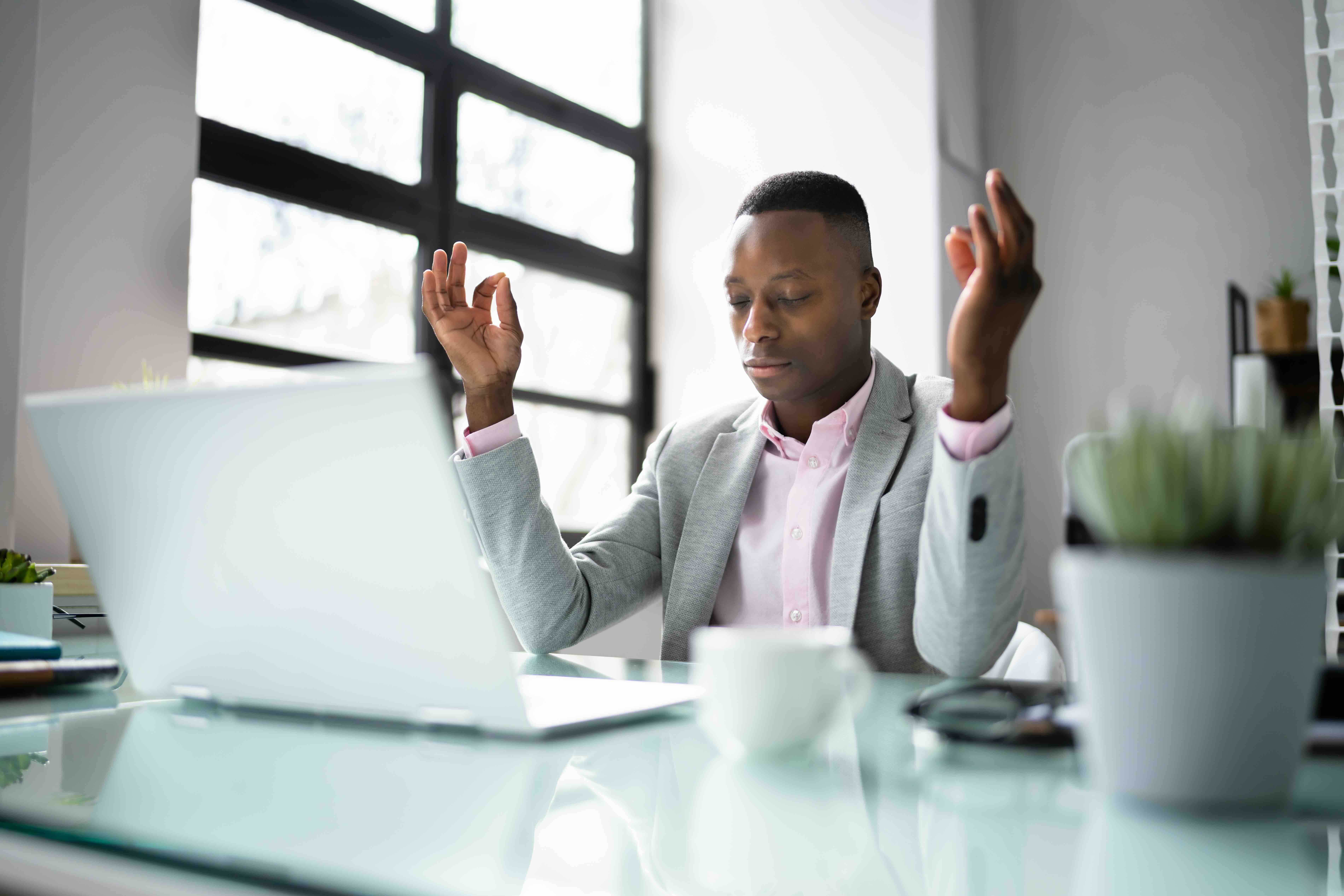 Professional man in gray blazer meditating at desk with laptop and coffee in bright modern office