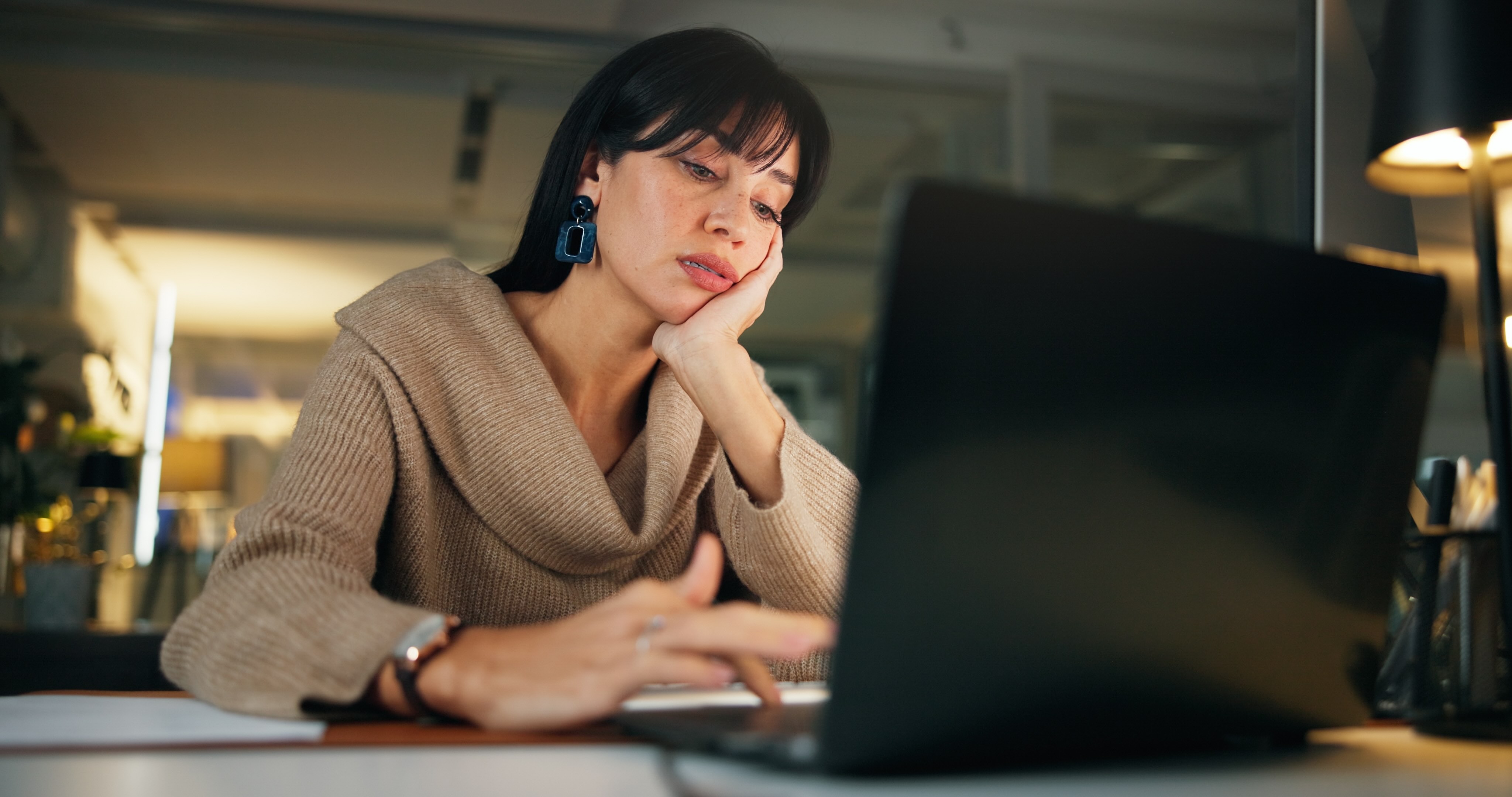 Woman looking exhausted while working at laptop, resting head on hand showing early signs of burnout