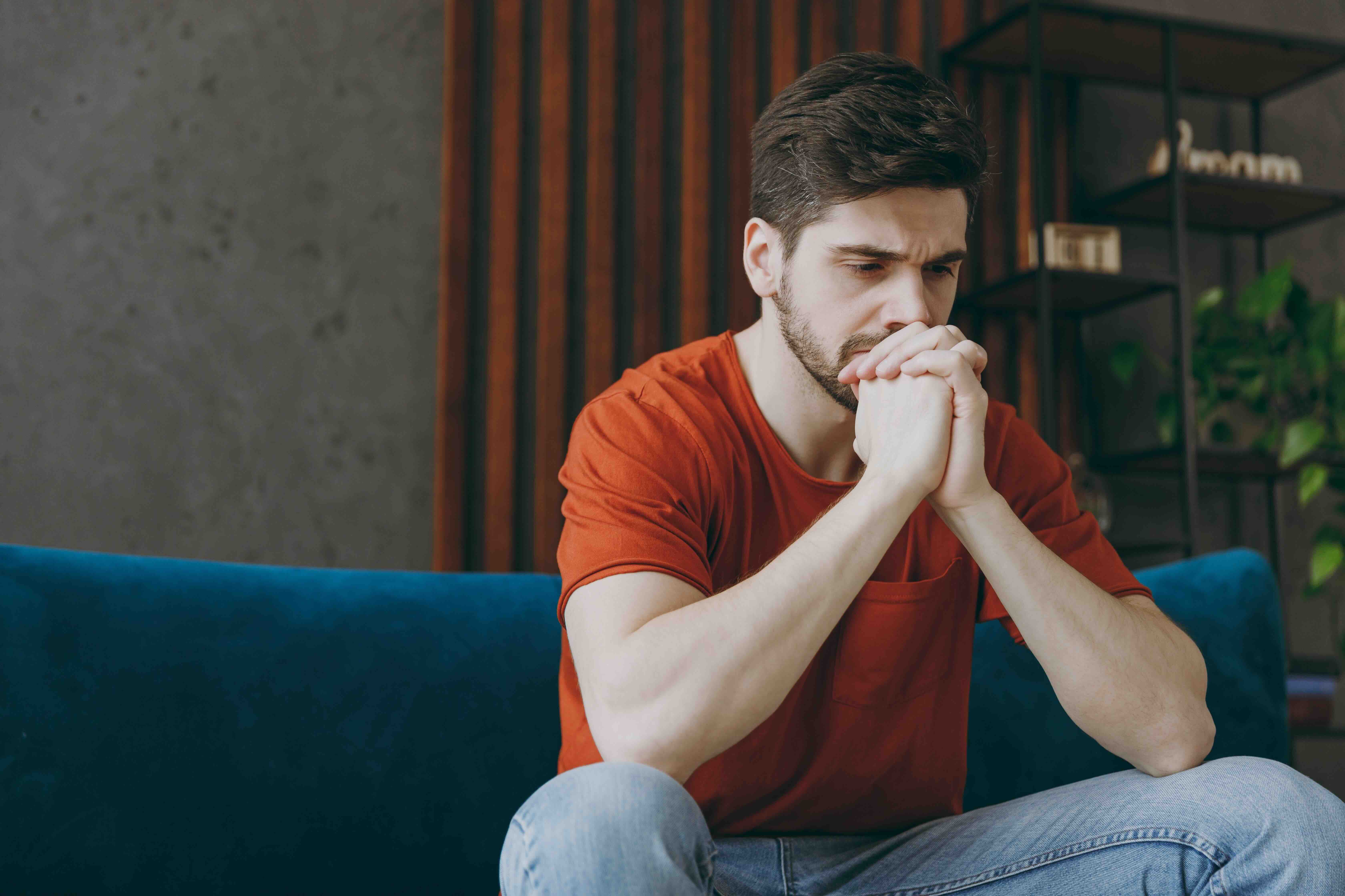 Man in orange shirt sitting on blue couch with hands clasped, looking worried and contemplative