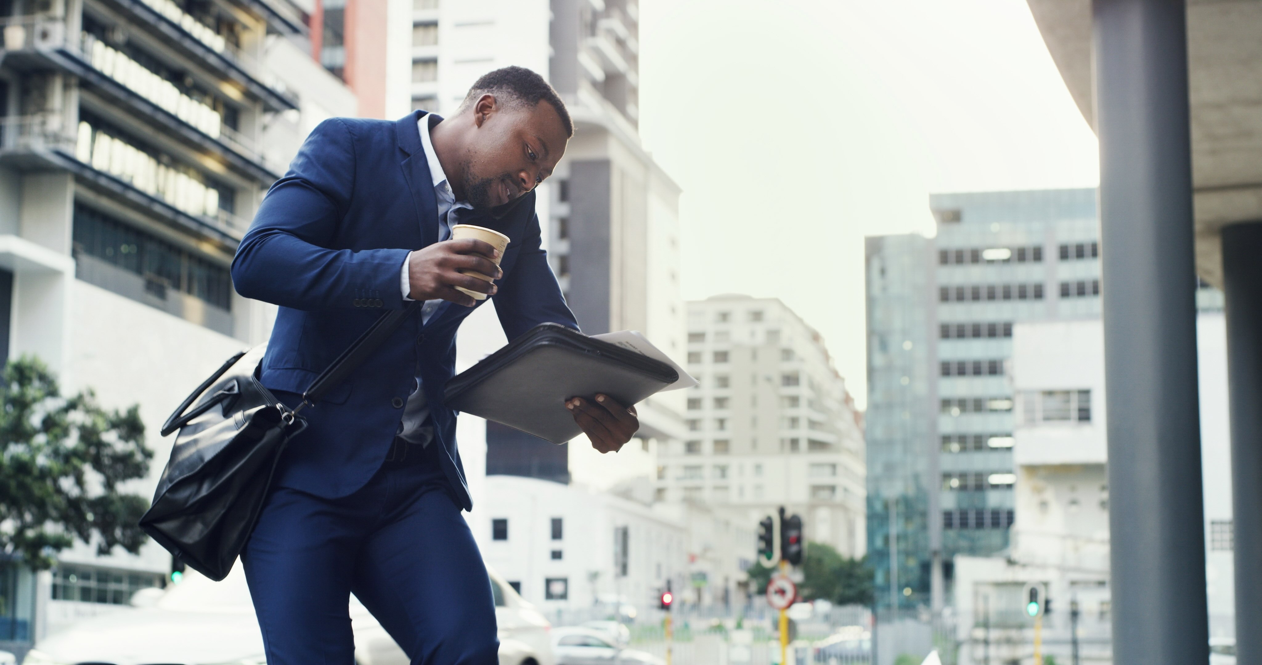 Businessman in navy suit multitasking on city street, holding coffee and tablet while rushing between tasks