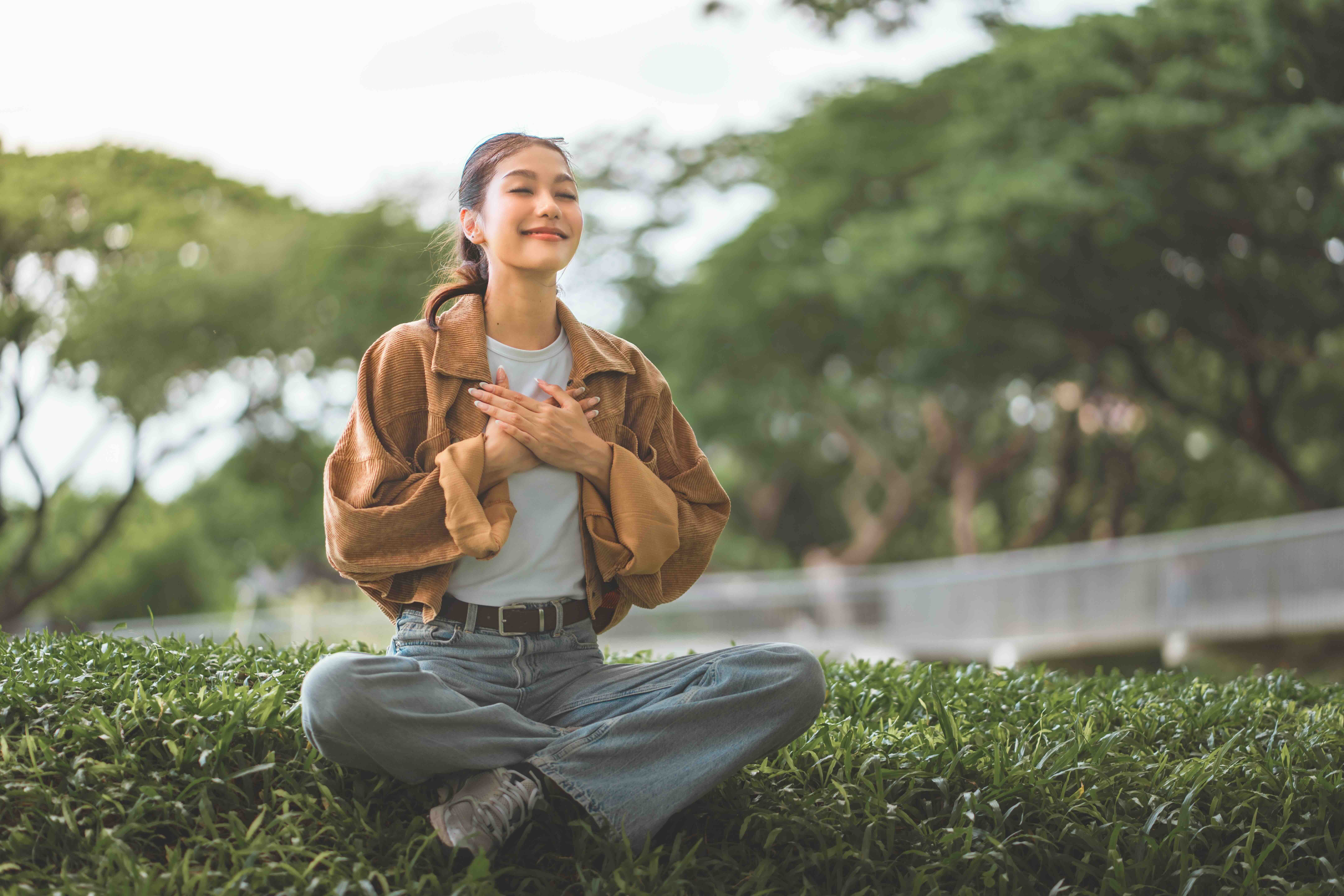 Woman meditating outdoors, hands over heart, smiling with eyes closed amid trees and green grass.