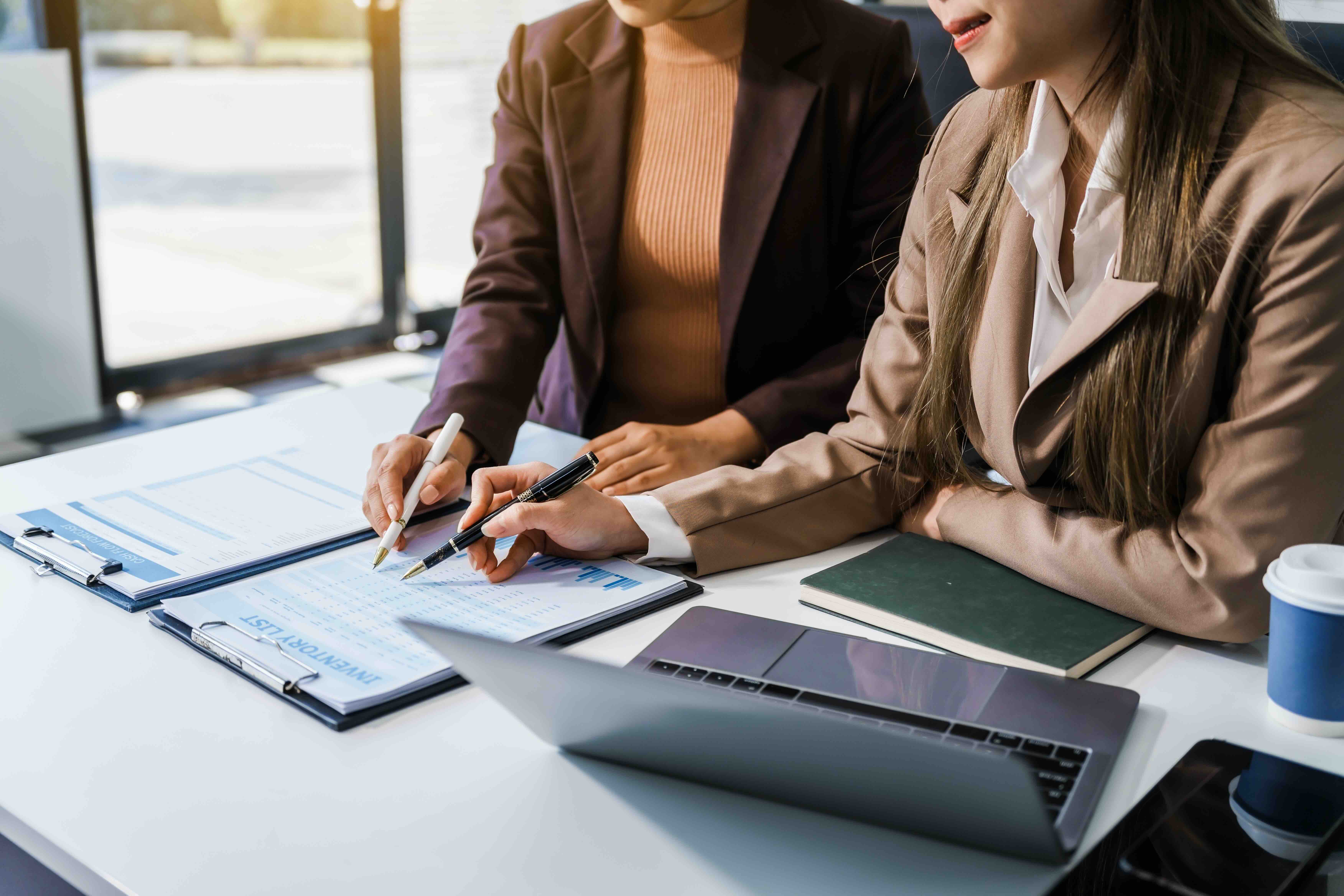 Two businesswomen reviewing financial charts and data at desk with laptop during collaborative work meeting
