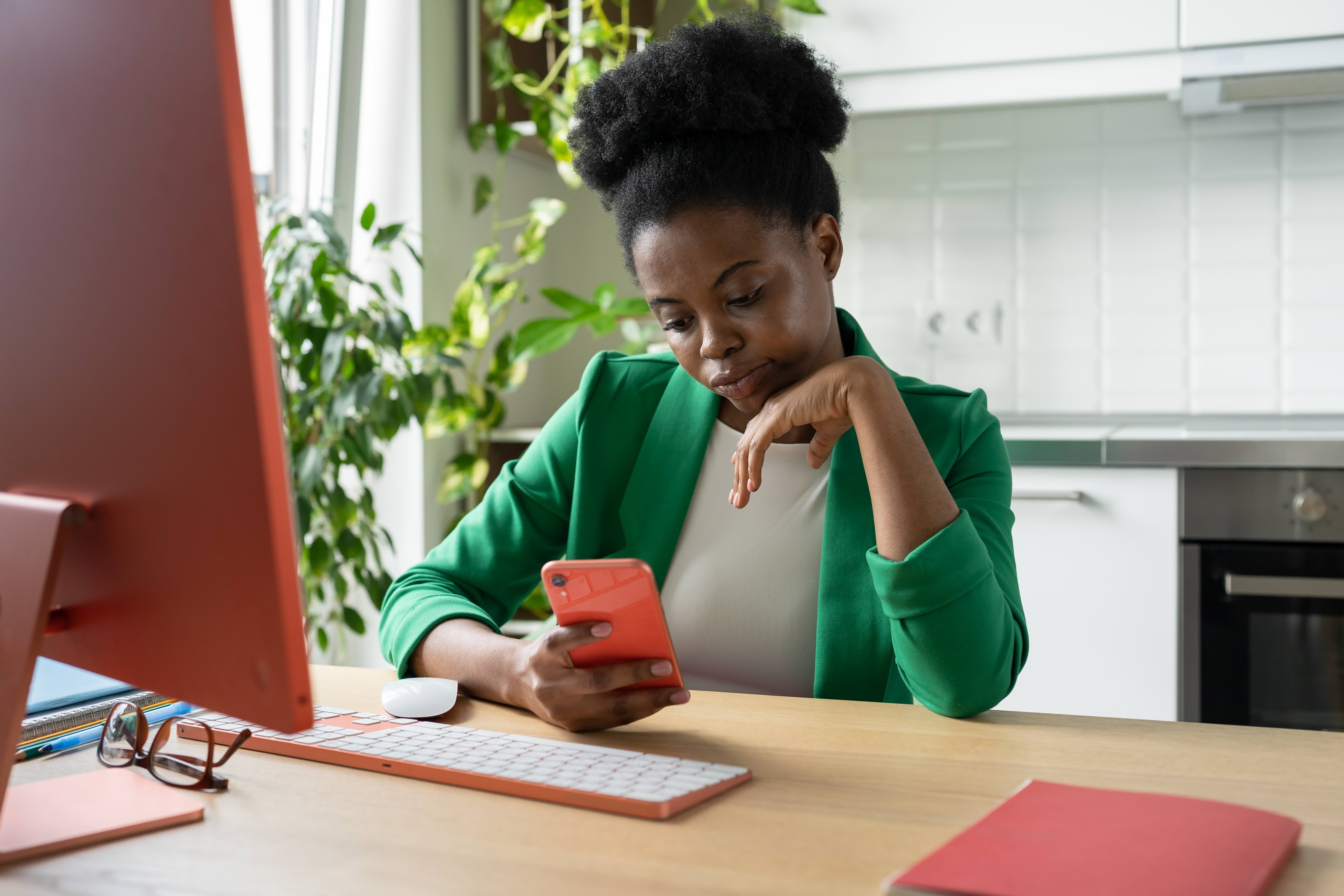 Woman in green blazer looking at phone while working at desk, appearing hesitant or contemplative